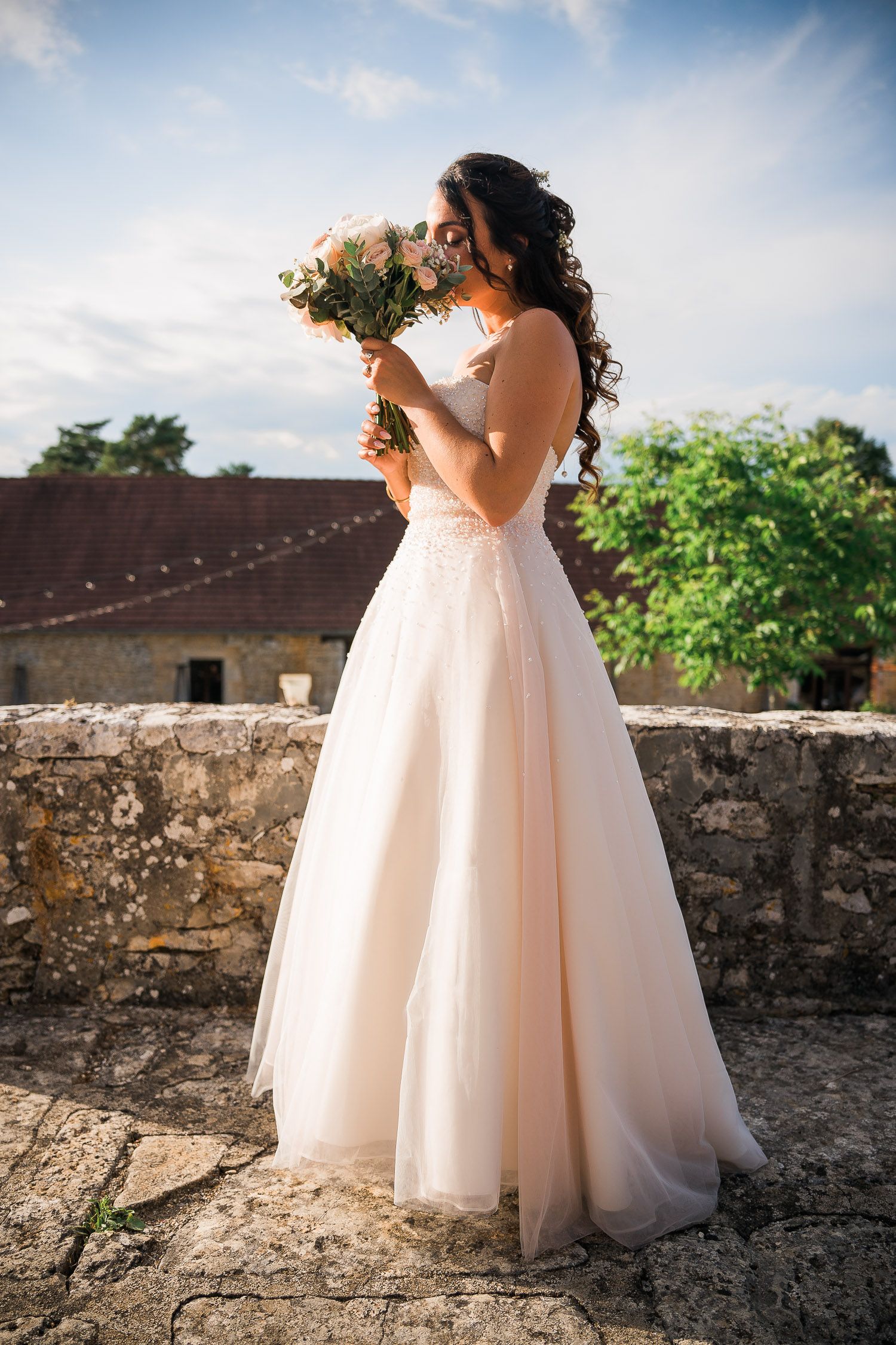 Bride in flowing blush gown posing against stone wall outdoors.