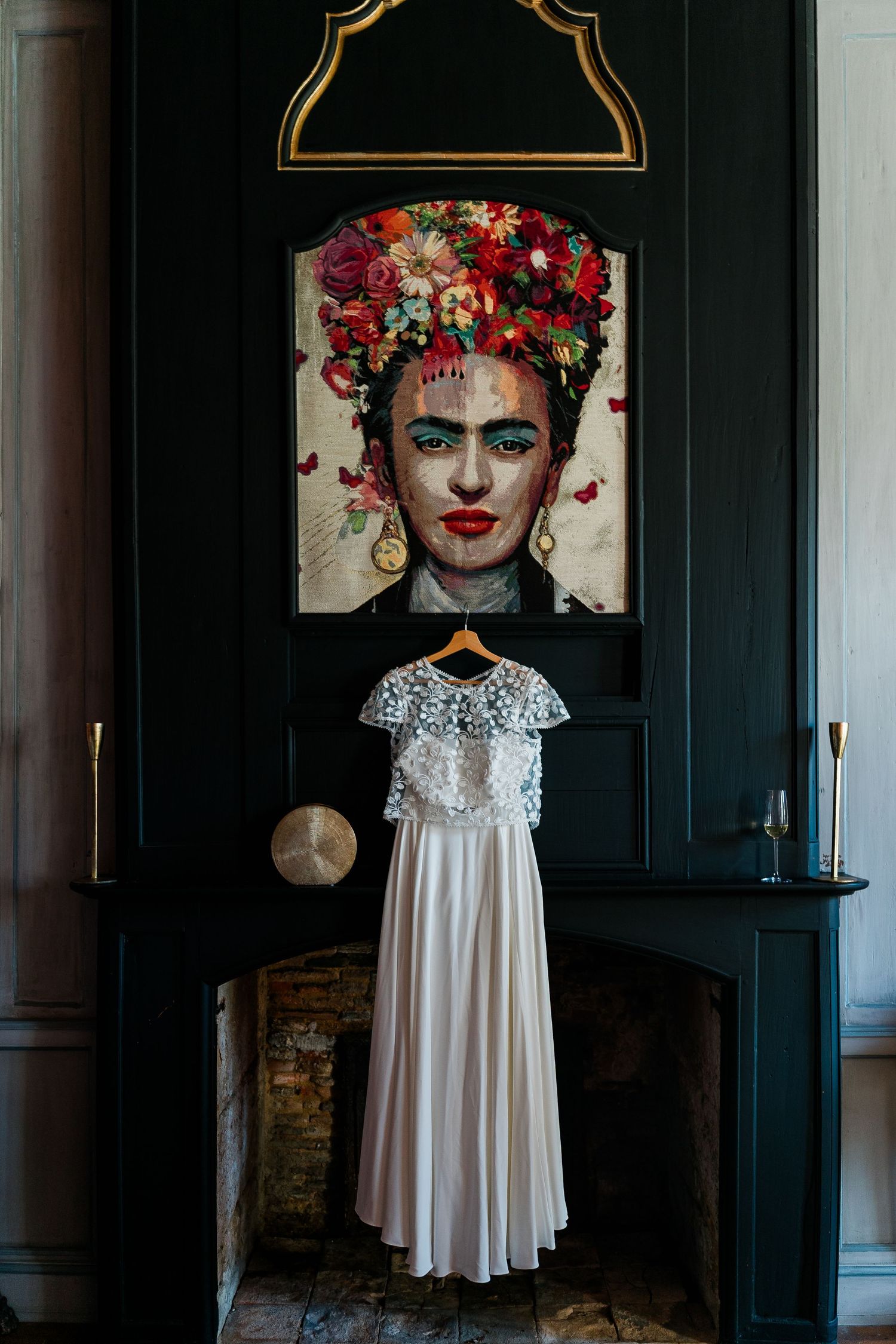 White lace wedding dress hanging beneath artistic portrait in dark green room with fireplace.