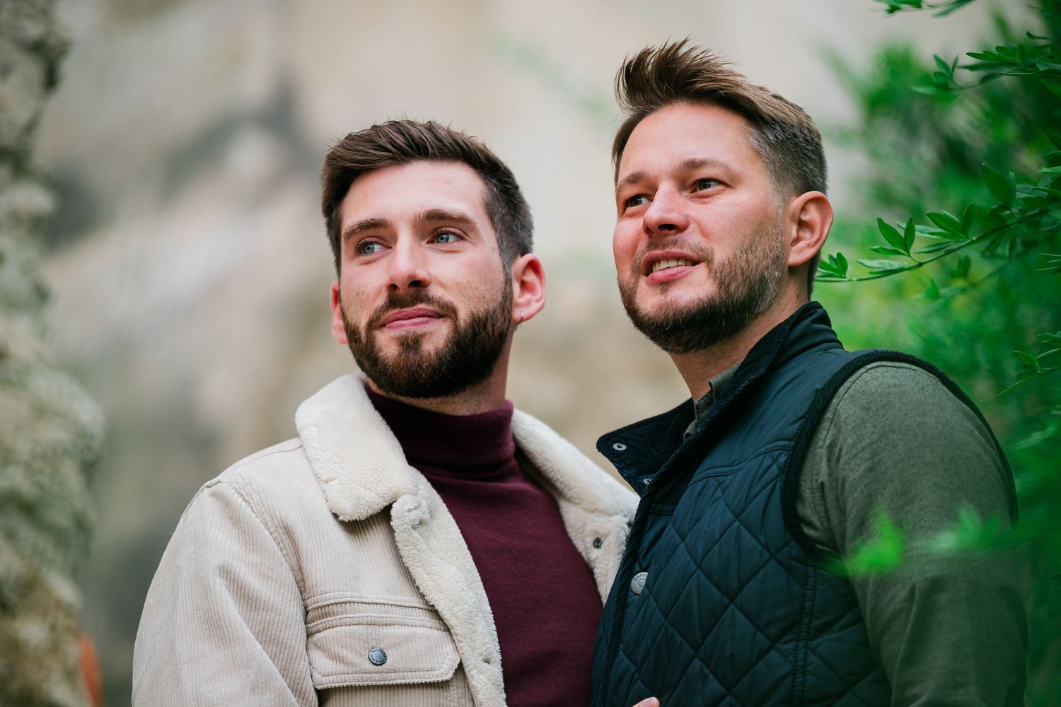Two people in casual outerwear pose together against stone wall background.