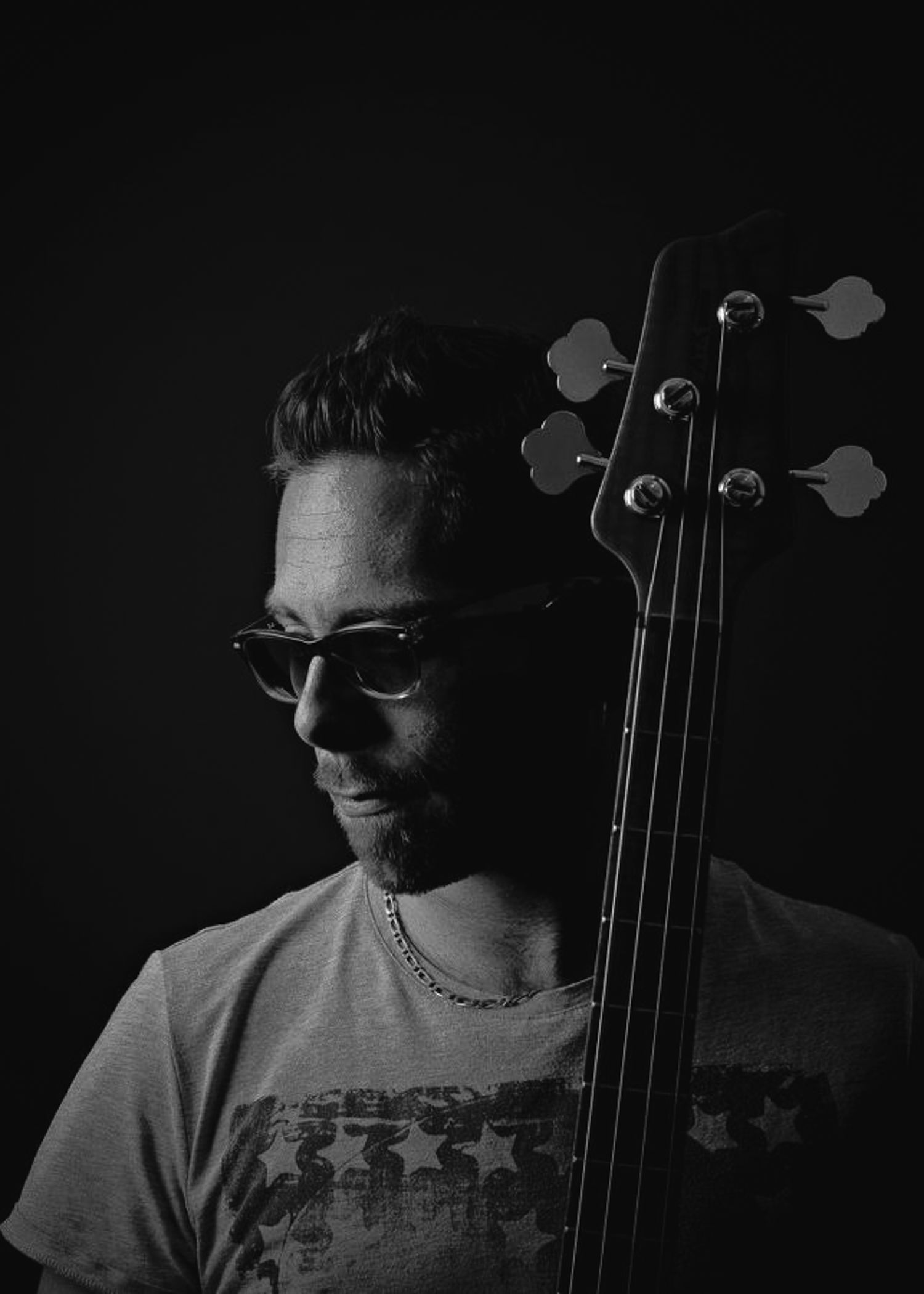 Black and white portrait of a musician with sunglasses holding a bass guitar neck against dark background.