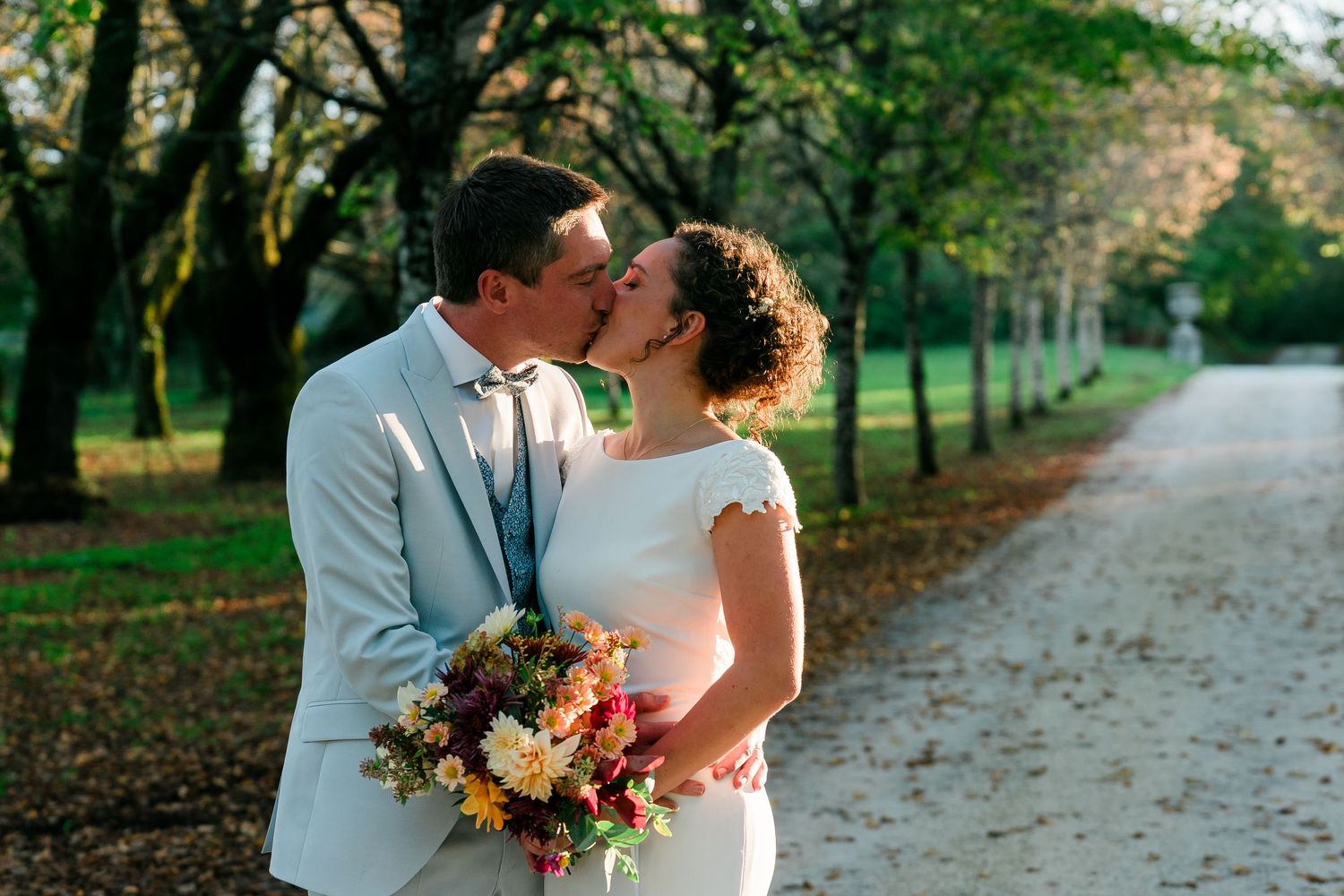 Newlyweds share a romantic kiss on a tree-lined path while holding a floral bouquet.