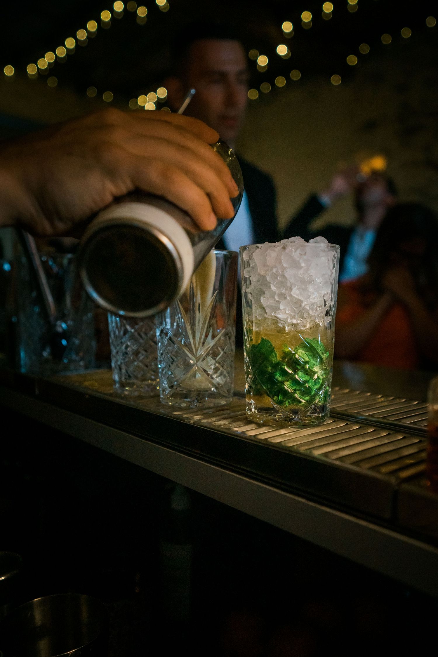 Bartender pouring cocktail into crystal glass with ice and green liquid.