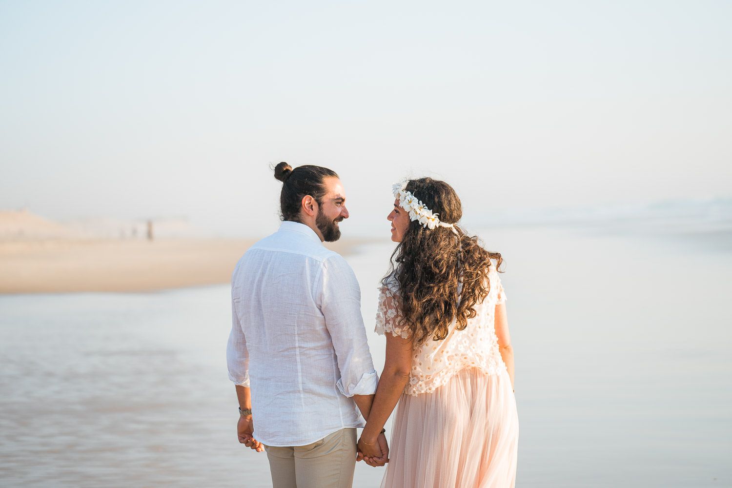 A romantic couple walks hand in hand along a misty beach at sunset wearing coordinated white and peach outfits.