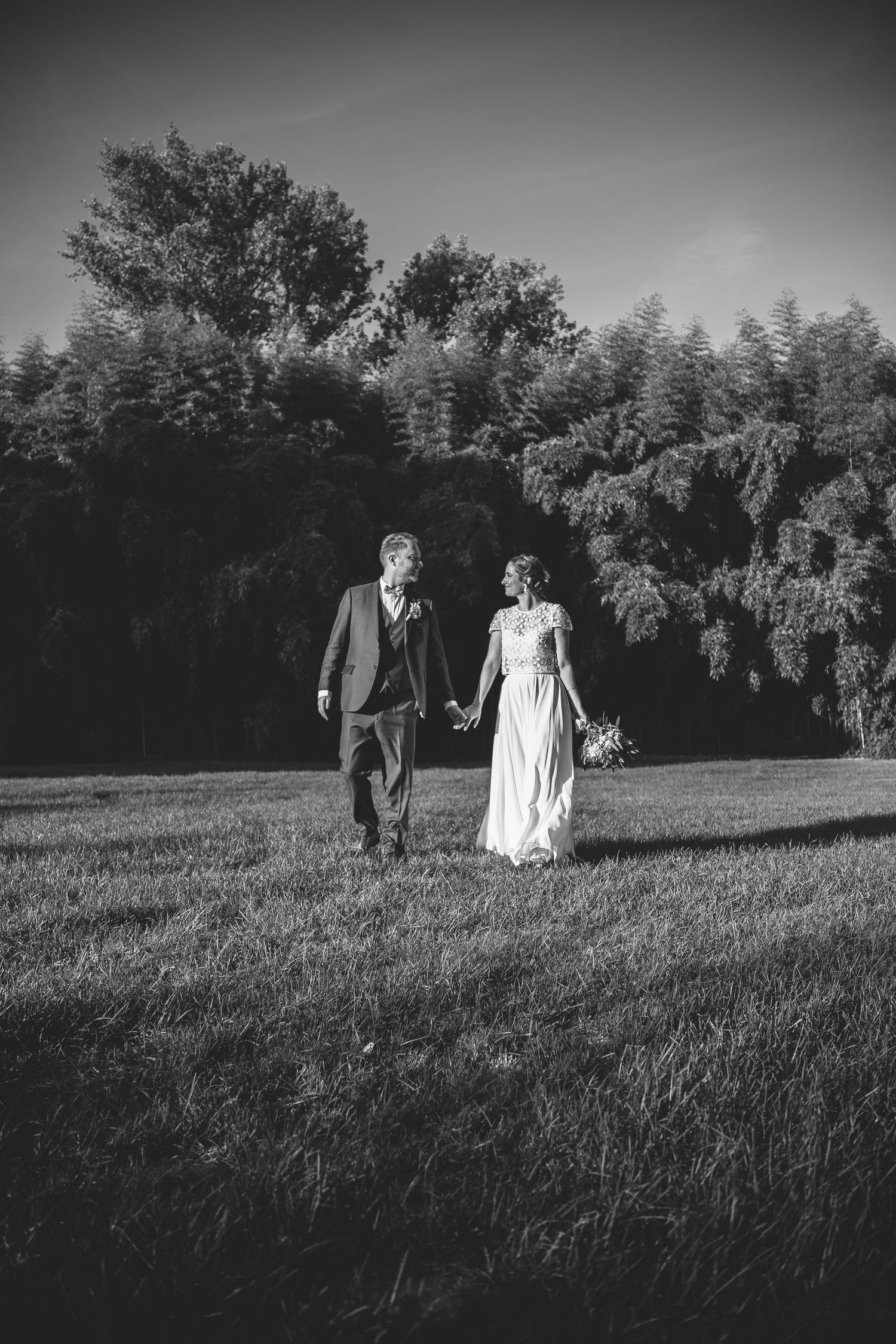 Black and white photo of a couple walking through a grassy field with trees in the background.