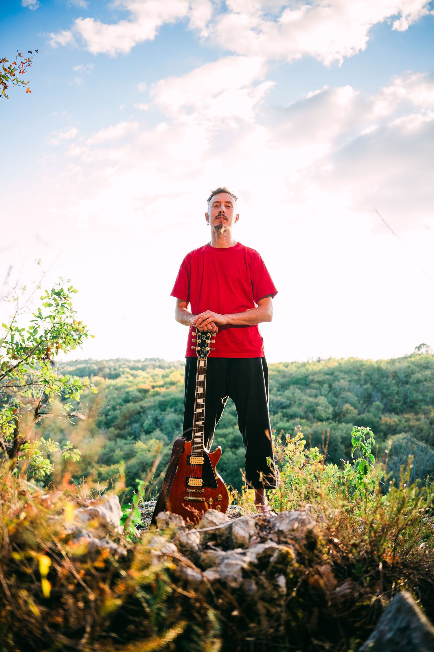 Musician stands on hilltop at sunset holding an electric guitar with scenic natural landscape in background.