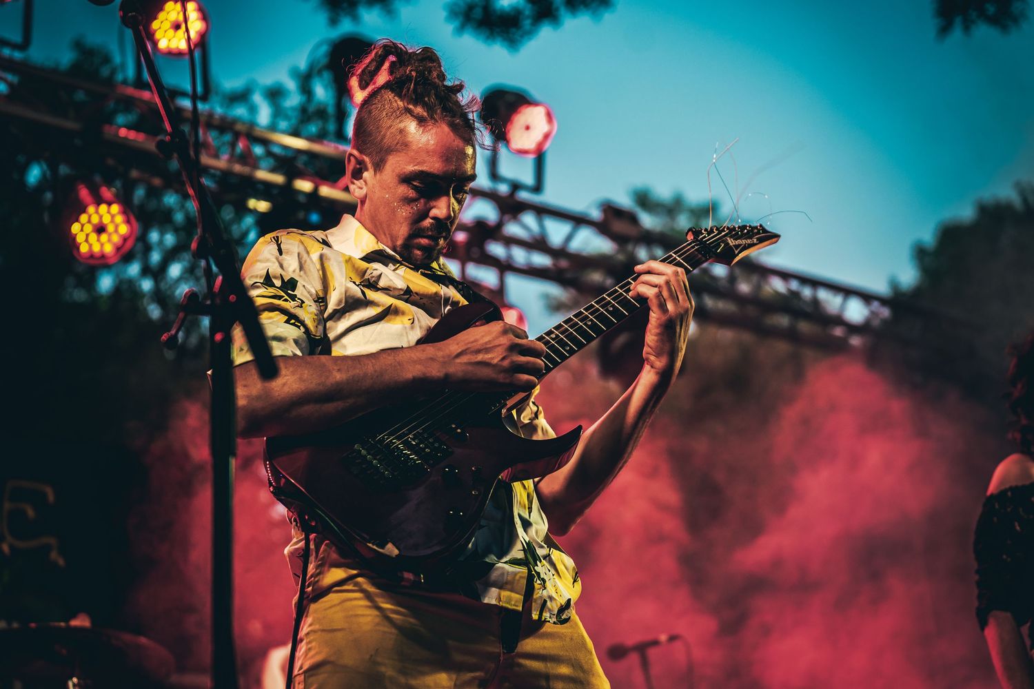 A musician performs on stage with a guitar amid dramatic blue and red lighting effects during an evening concert.