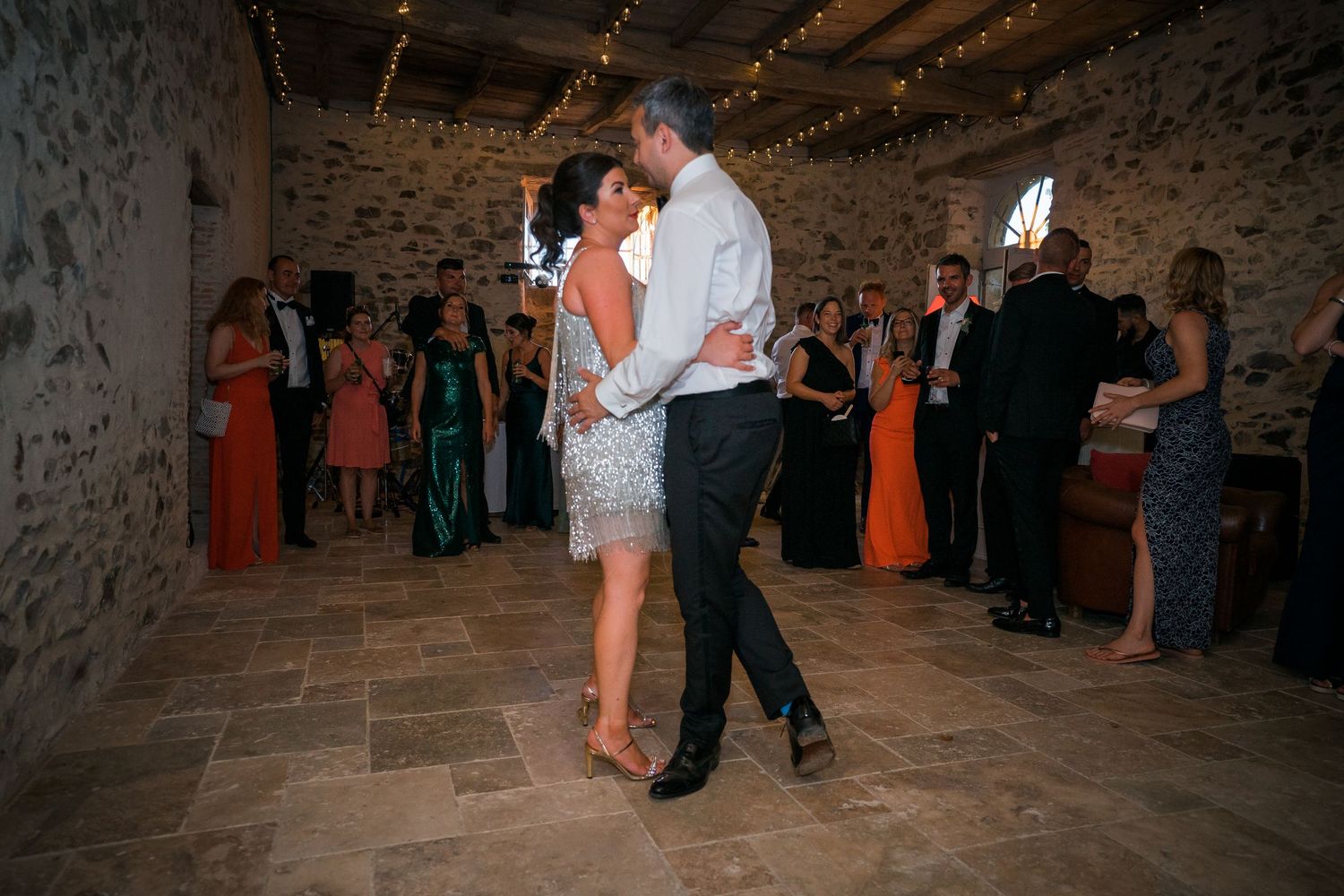 Dancing couple surrounded by guests at rustic stone venue with string lights.