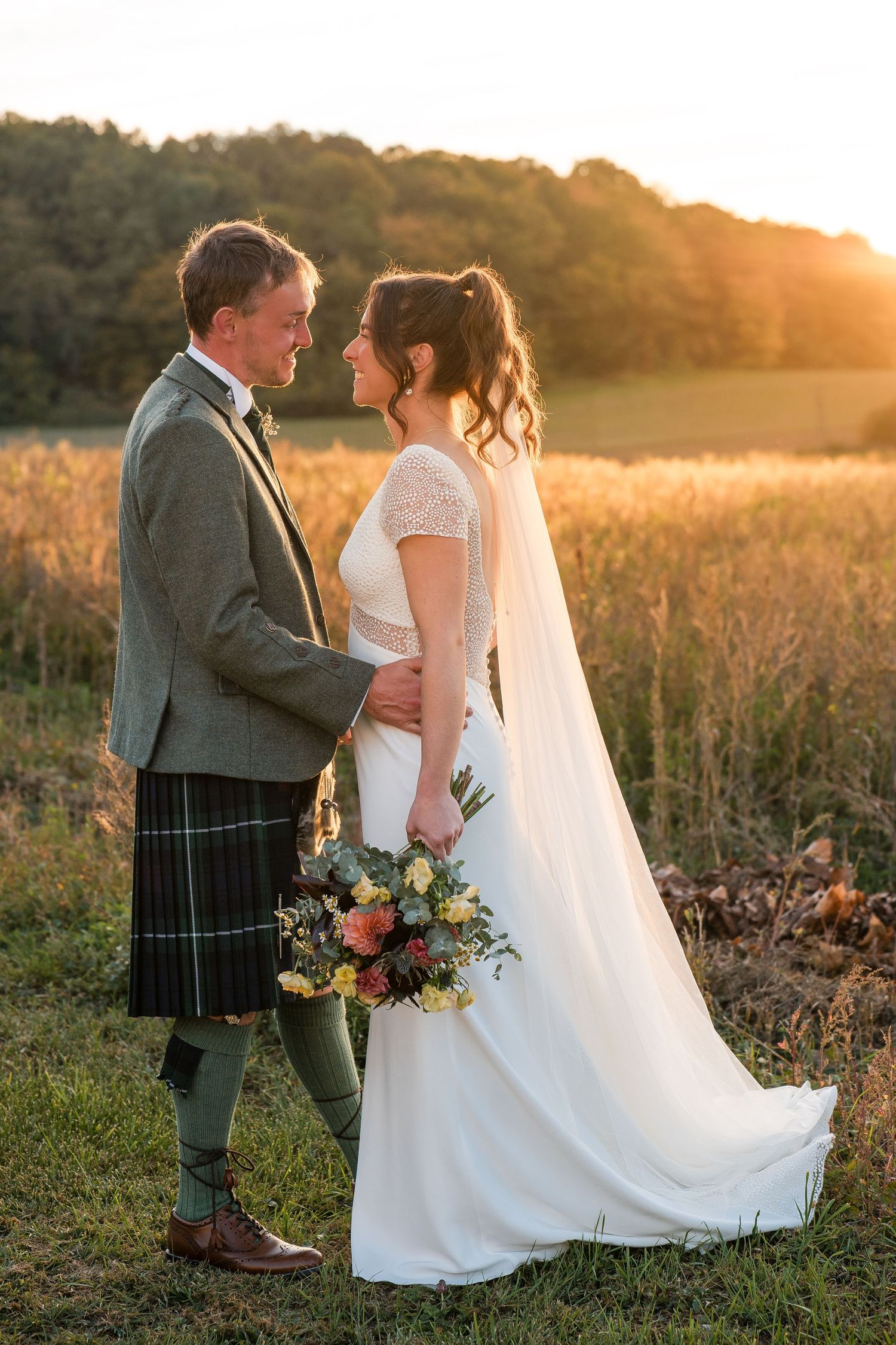 Sunset wedding portrait in field with bride in white dress and groom in traditional Scottish kilt.