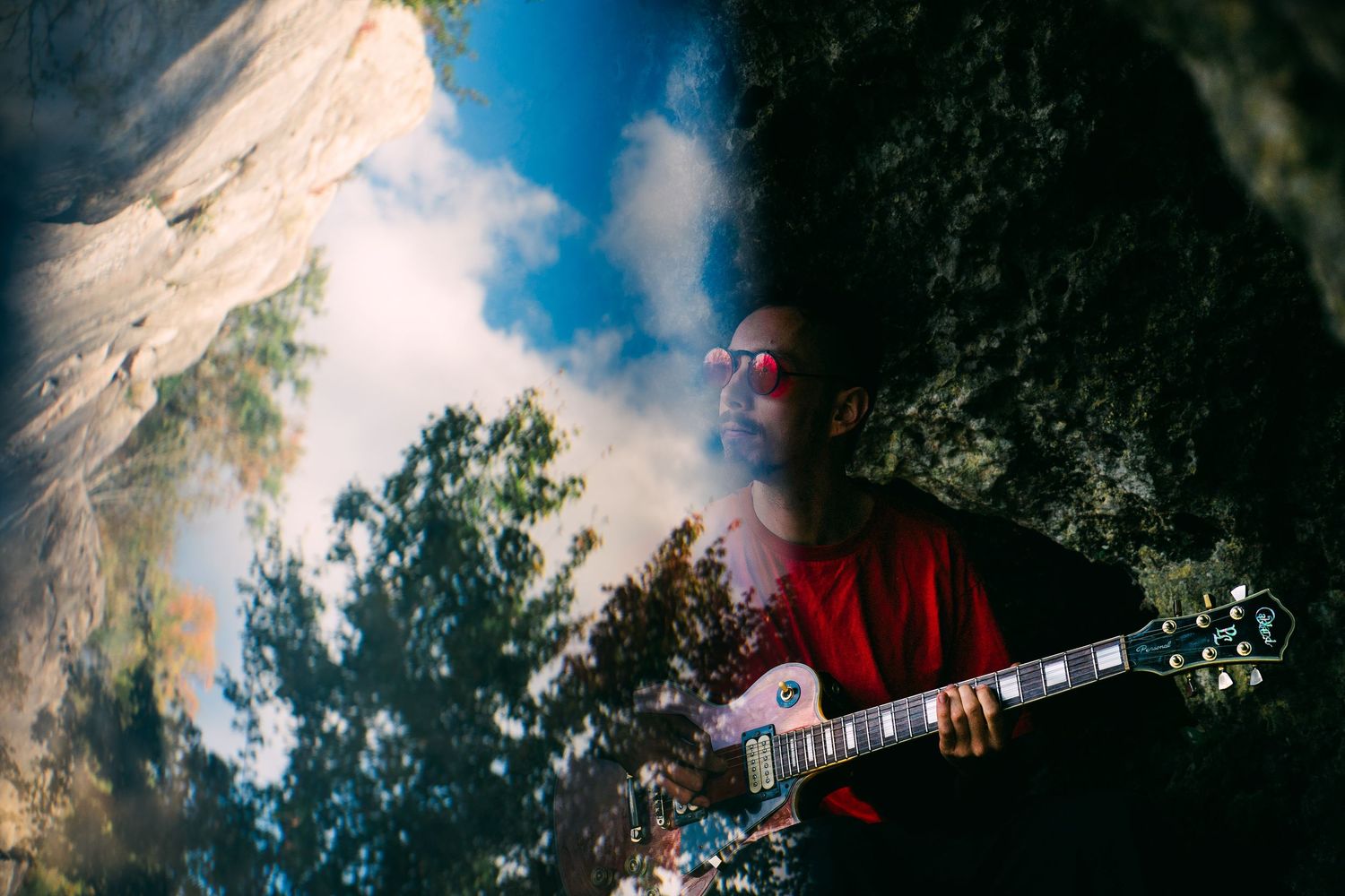 A guitarist plays an electric guitar outdoors against a dramatic sky with trees and clouds in the background.