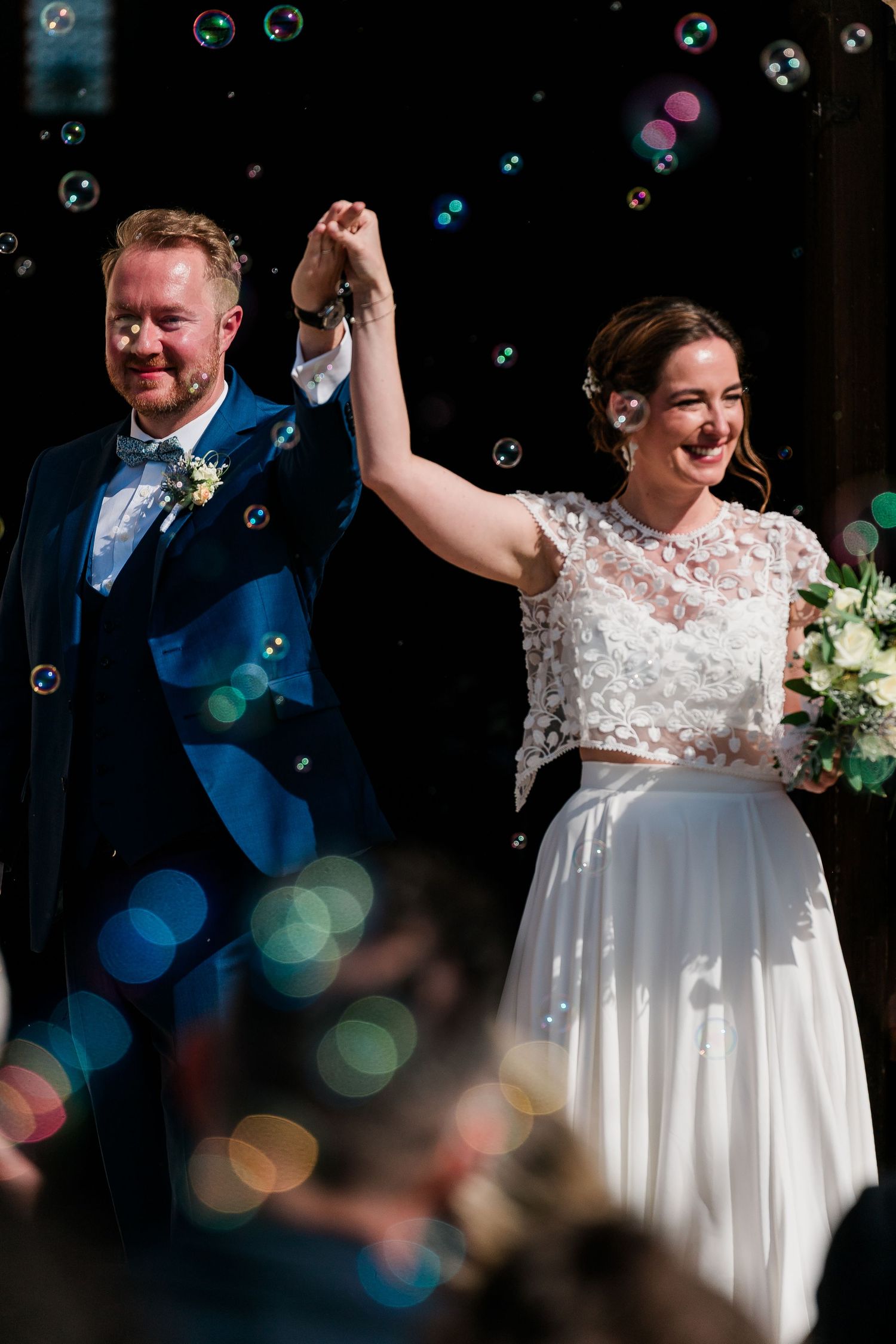 Newlyweds celebrate with raised hands amid floating bubbles and bokeh lights at their wedding celebration.