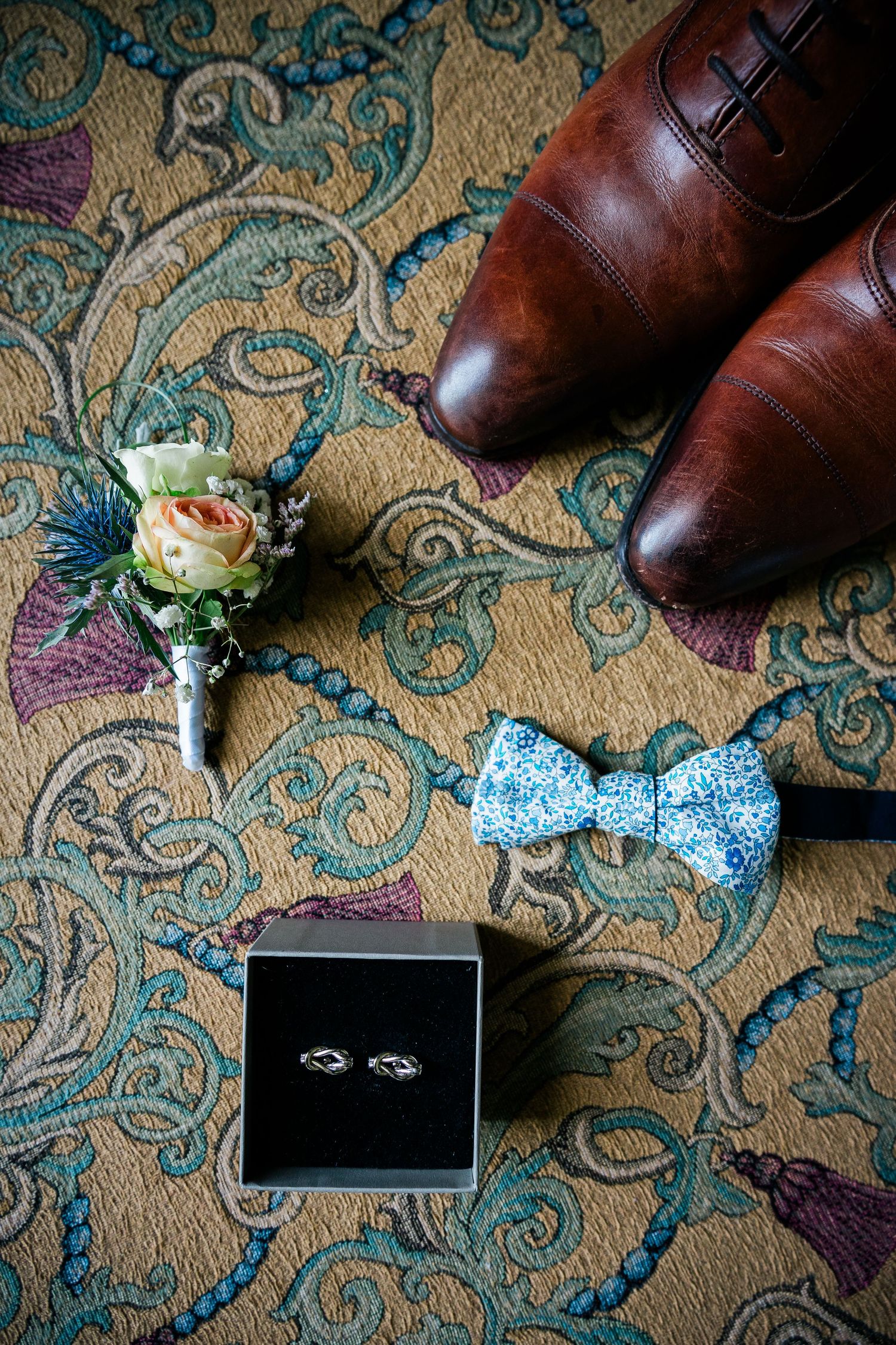 Wedding accessories including brown leather shoes, floral boutonniere, blue bowtie and wedding rings on ornate carpet.