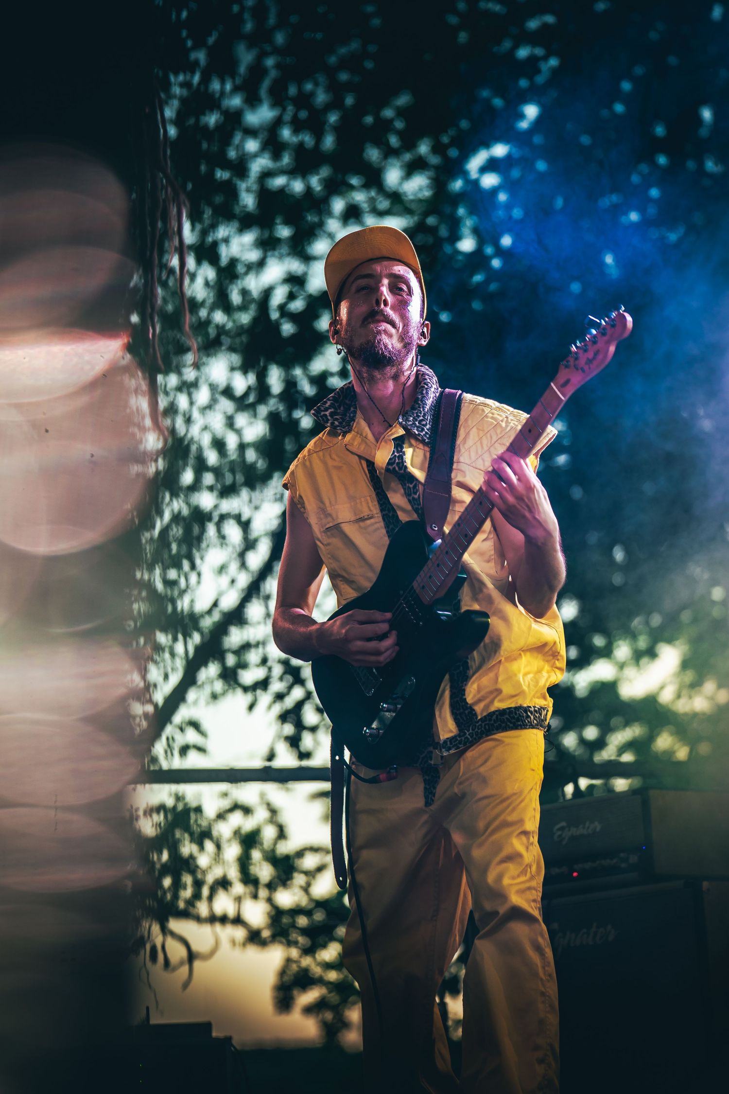 Musician performs on stage wearing yellow overalls and playing guitar against a moody blue-lit backdrop with trees.