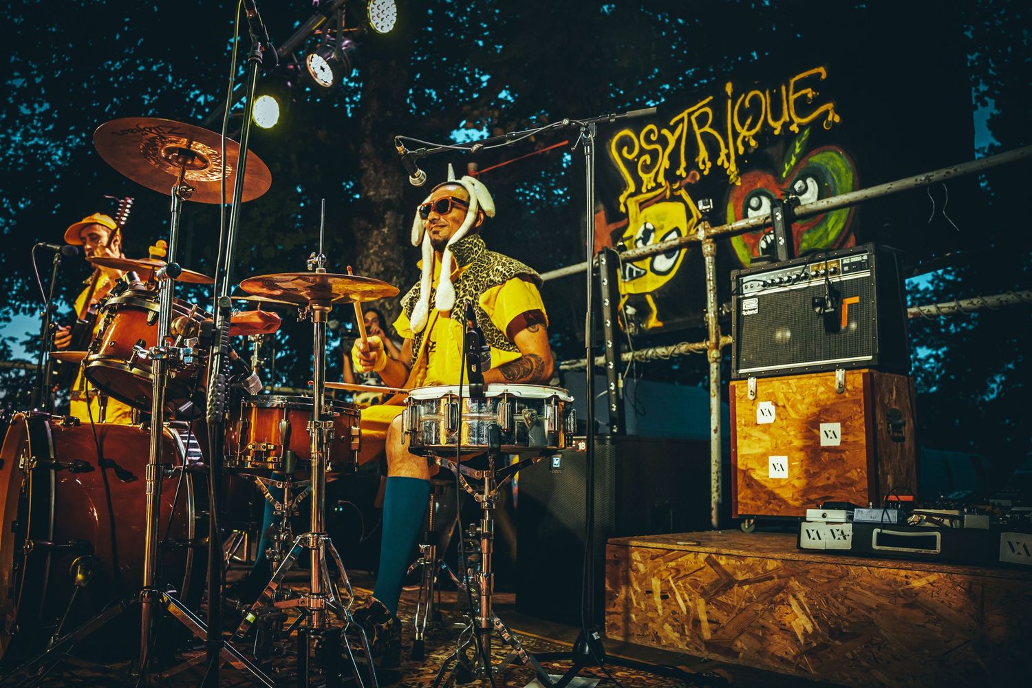 Band performs on a dimly lit stage with yellow lighting and musical equipment silhouetted against a dark backdrop.