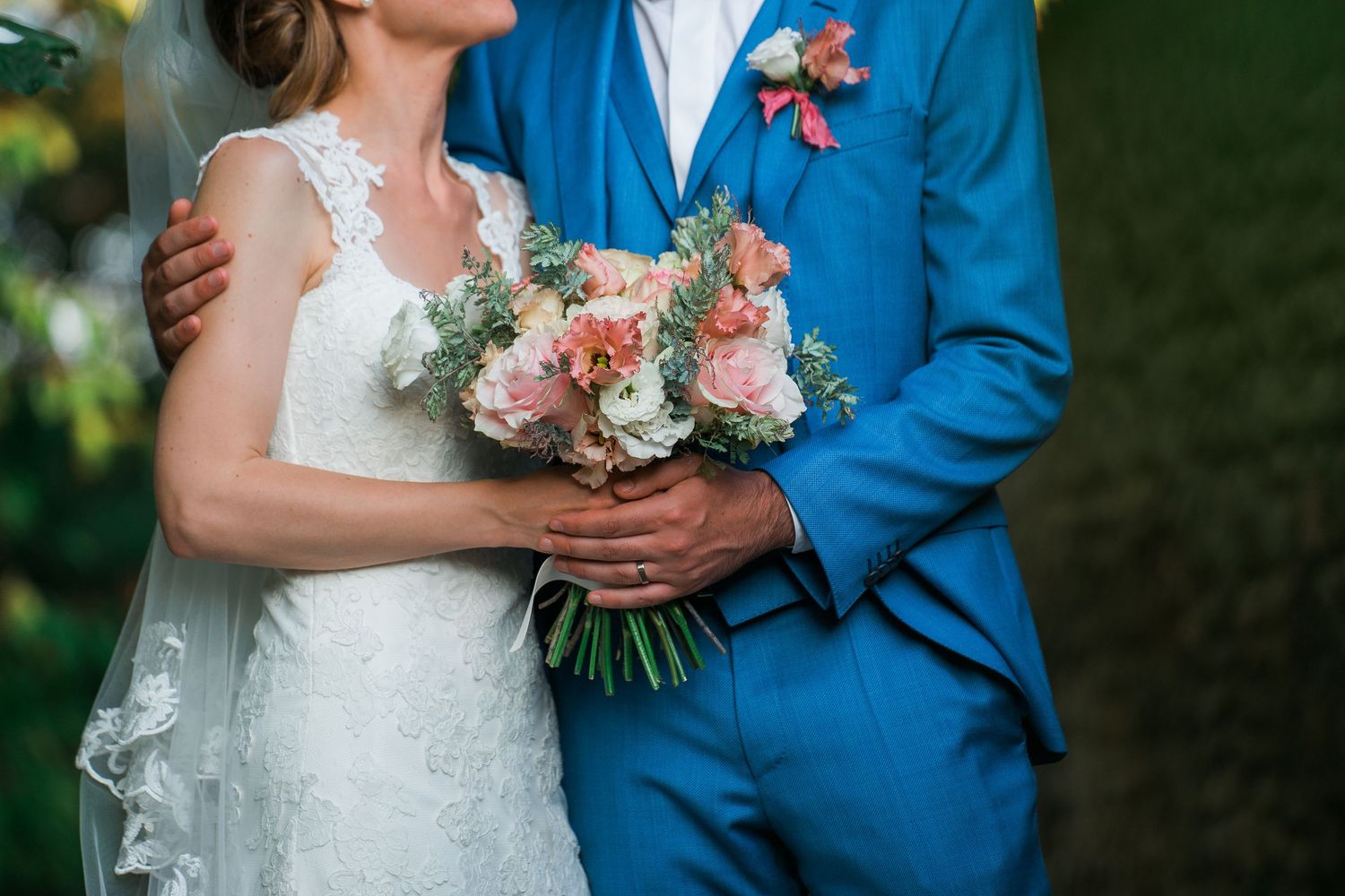 Wedding couple with bright bouquet poses against natural green background in formal attire.