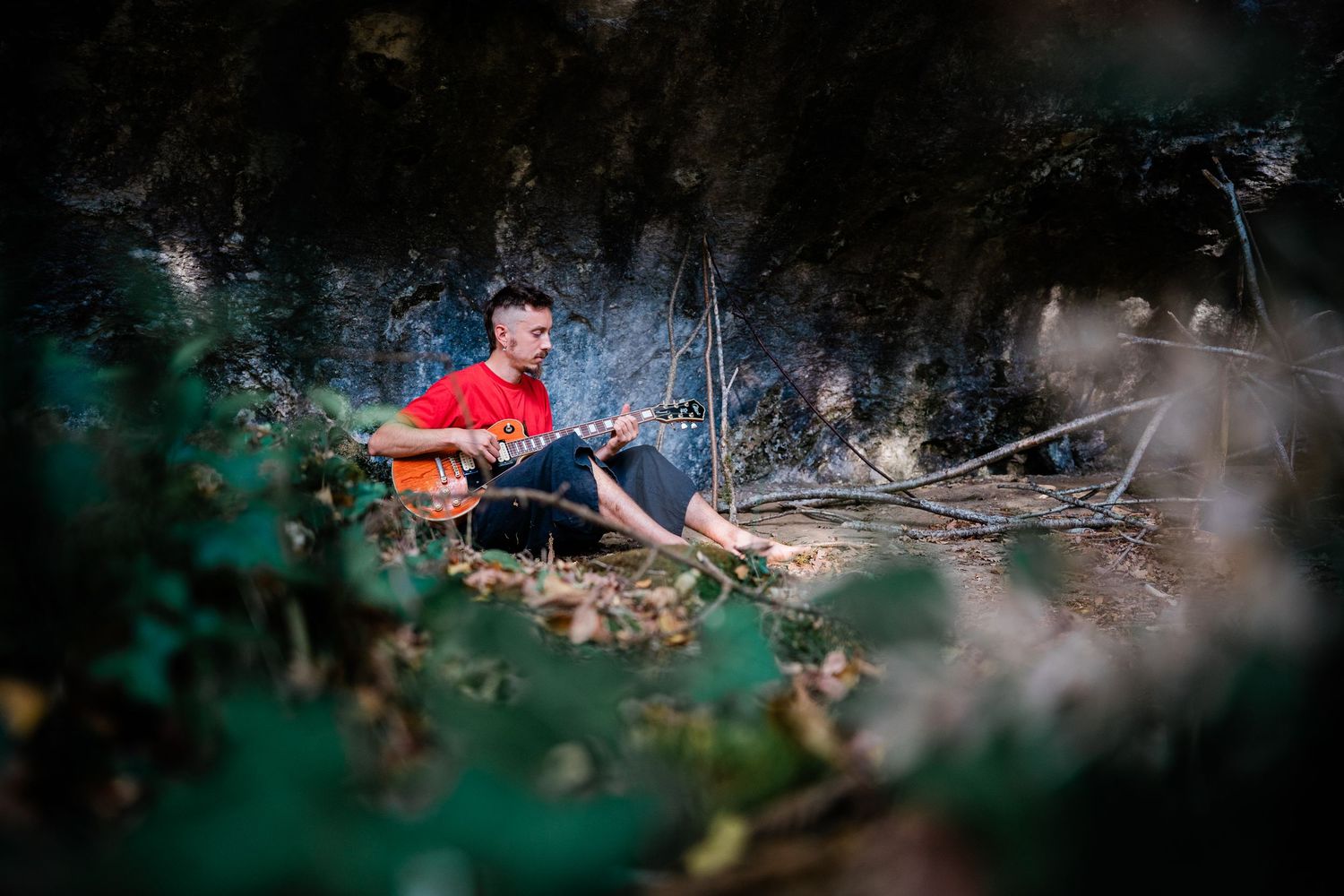 A person in a red jacket lies on a forest floor among scattered leaves and branches during an outdoor photoshoot.