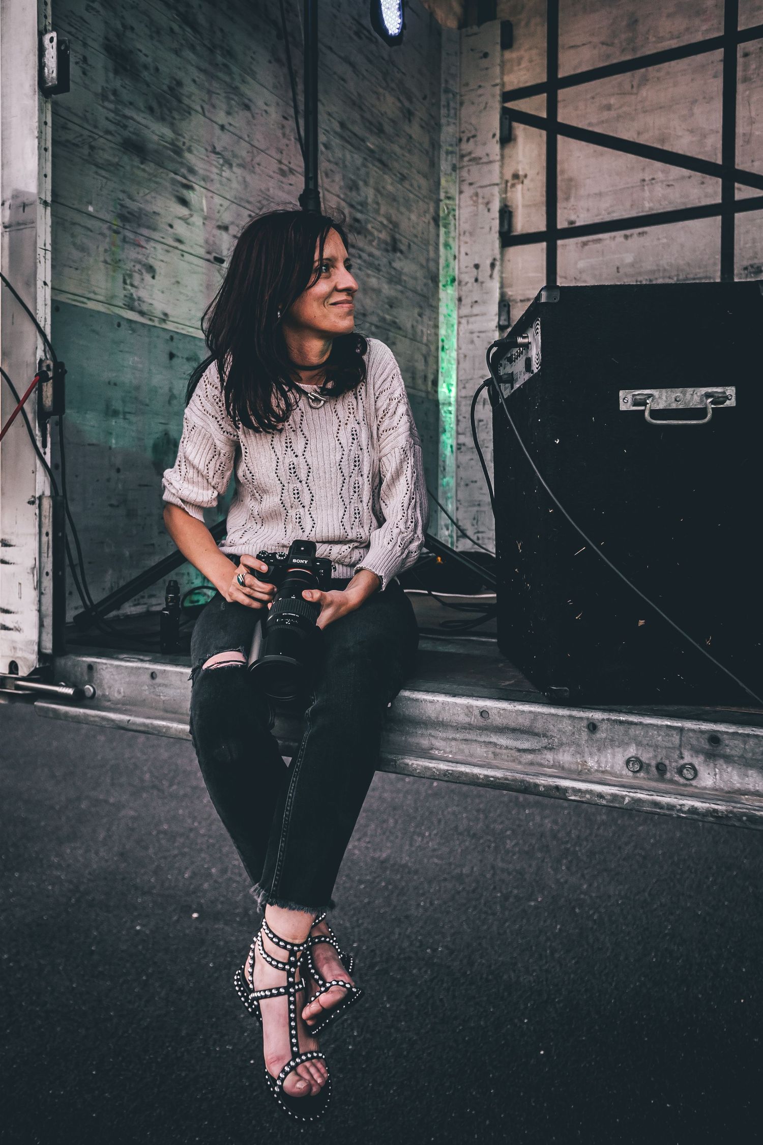 A person in casual attire sits in an industrial setting while holding a camera, creating a moody urban photoshoot vibe.
