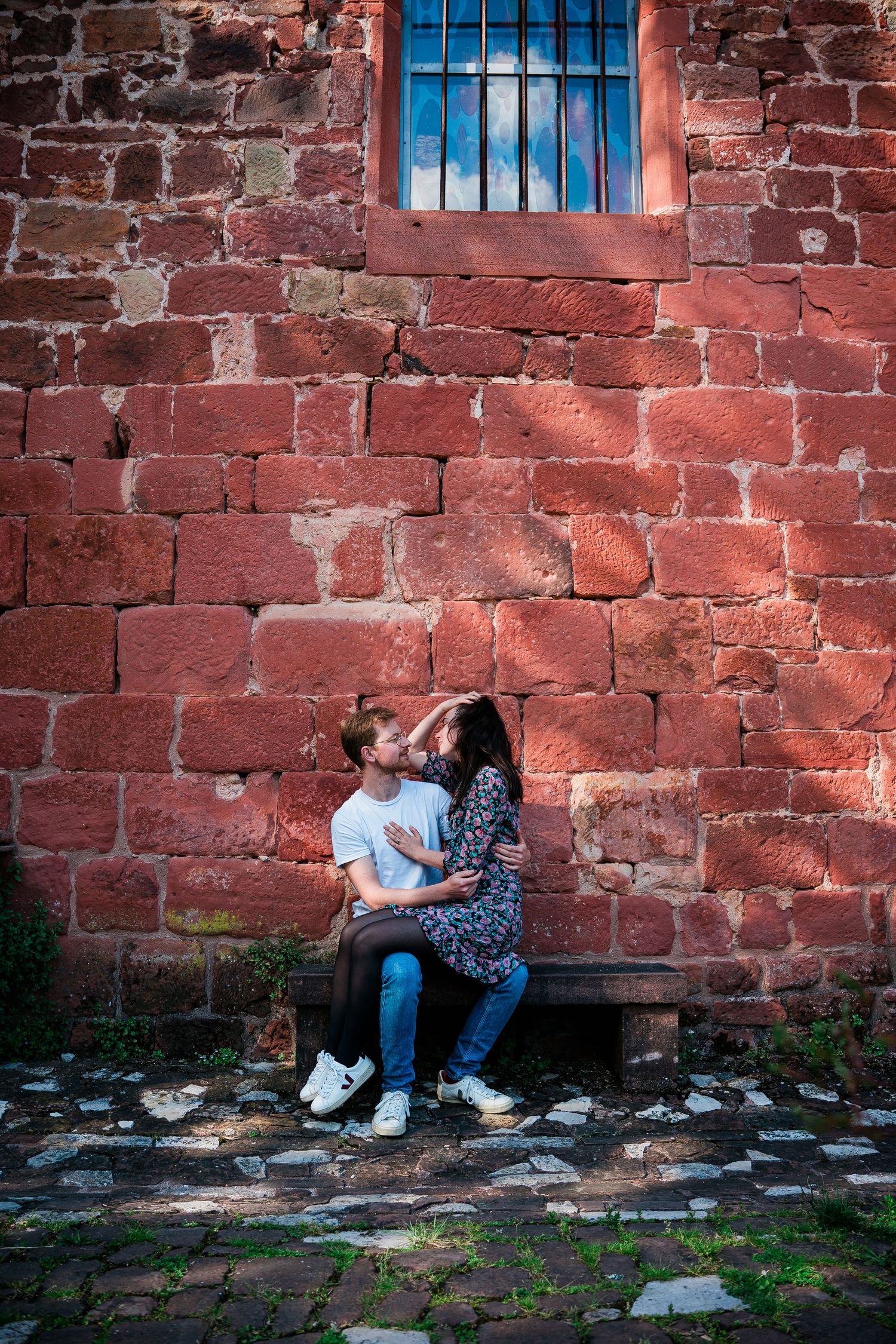 Couple sharing tender moment on bench against rustic red brick wall with barred window.