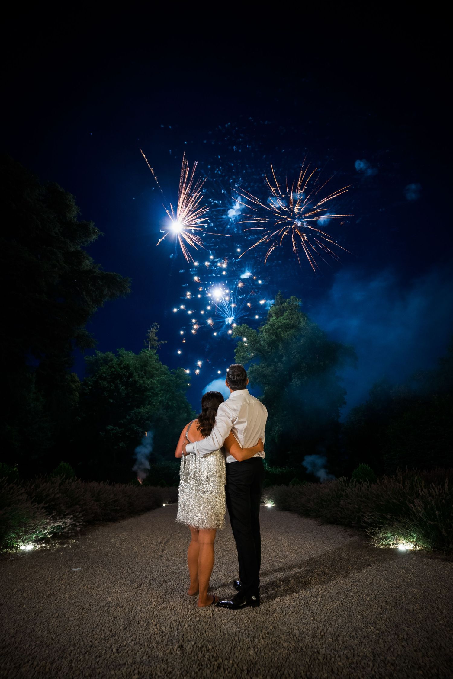 Couple watches fireworks display against dark blue night sky on garden path.