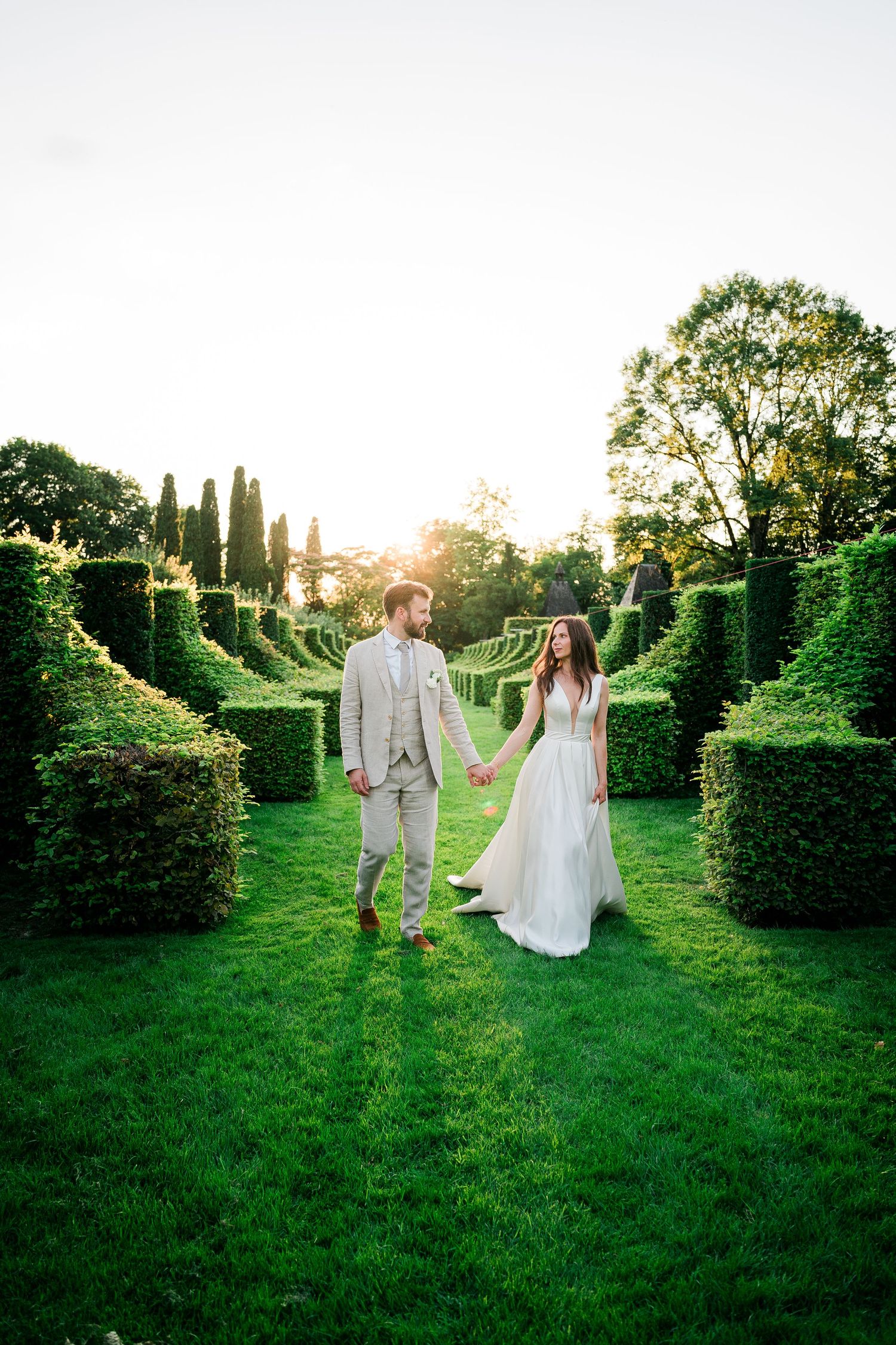 Couple walking through a lush garden with manicured hedges at sunset.