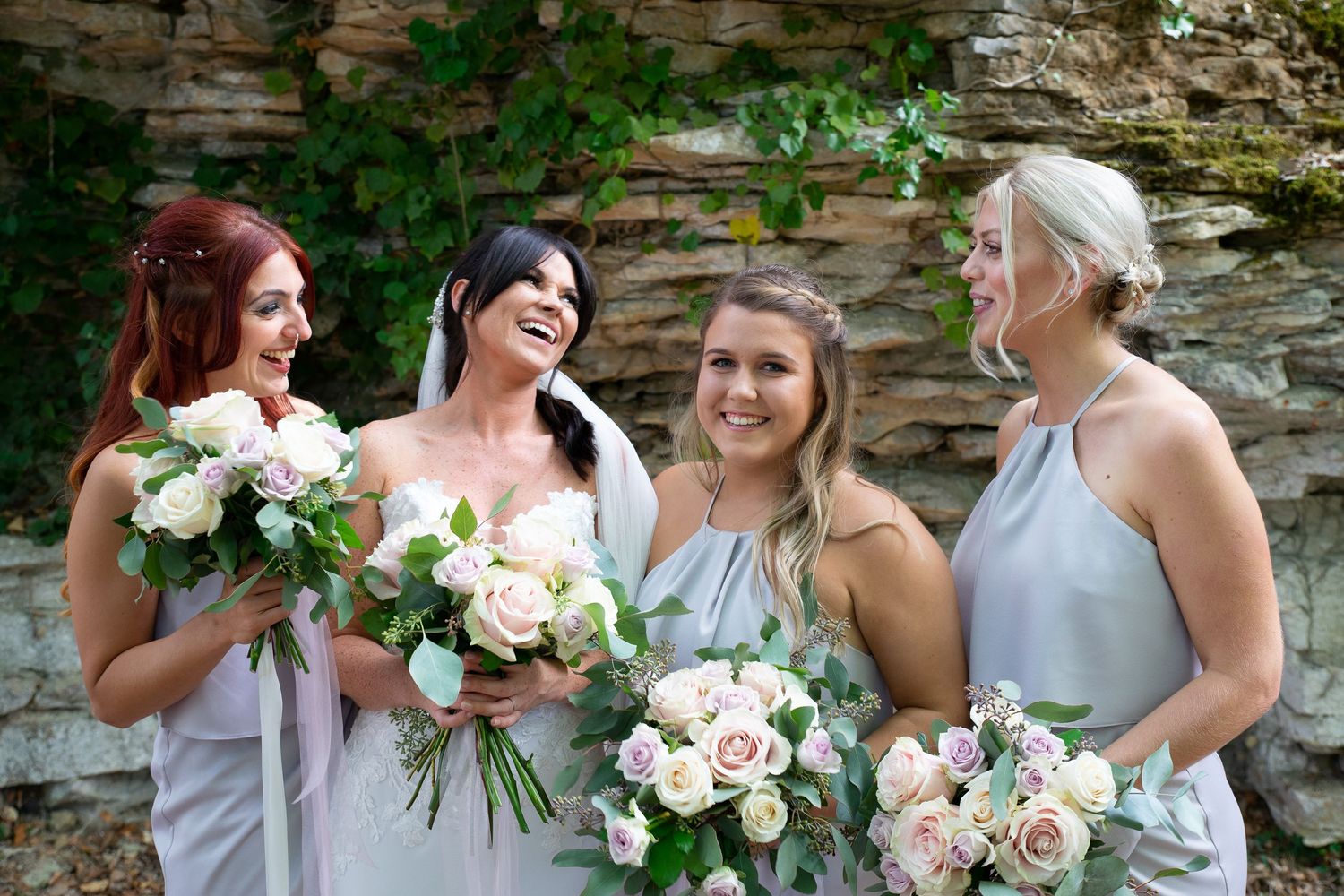 A group of bridesmaids in light colored dresses share a joyful moment against a stone wall backdrop.