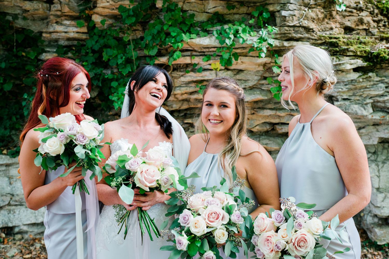 Bridesmaids in light gray dresses share joyful laughter while holding pink and white bouquets.