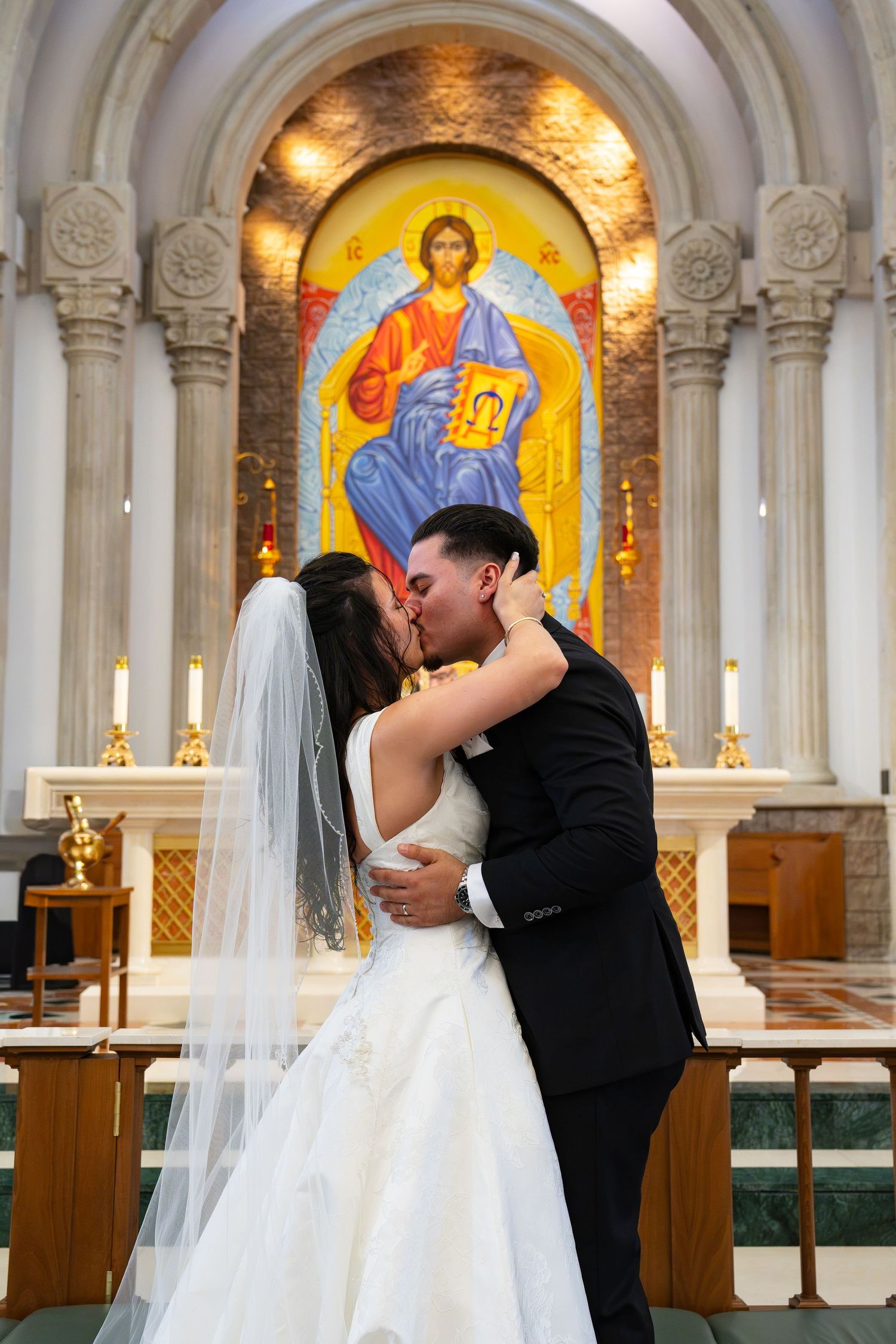 Bride and groom share first kiss at church altar with colorful Byzantine mosaic of Christ behind them.