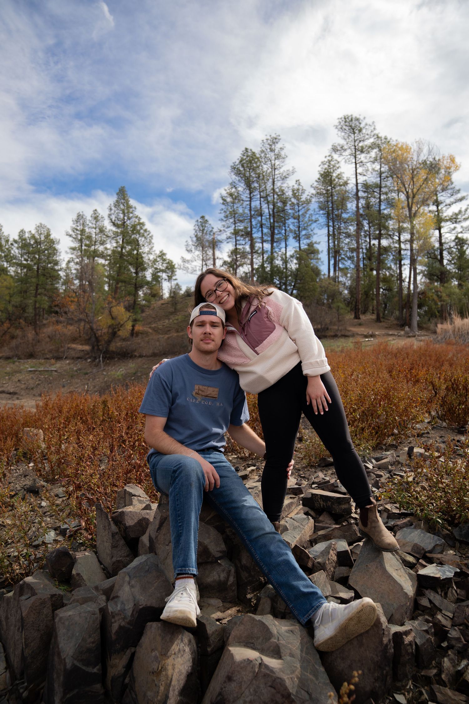 Couple relaxes on rocky outcrop with autumn forest and cloudy sky behind them.