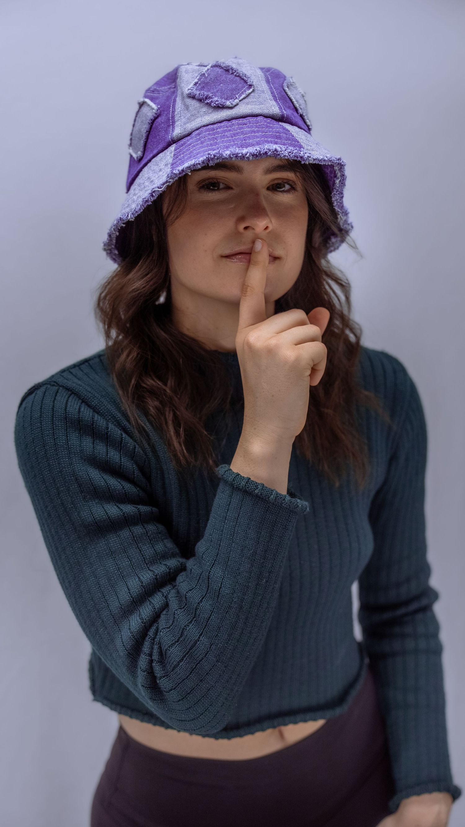Person in dark sweater and purple bucket hat making playful shushing gesture against light background.