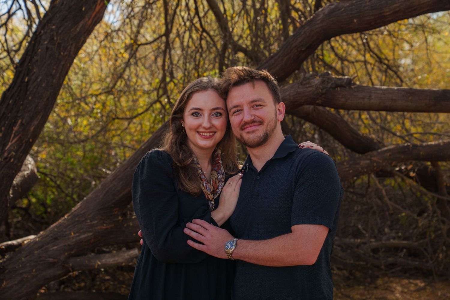 Photo of a couple posing together under autumn trees with golden yellow foliage in a park setting.