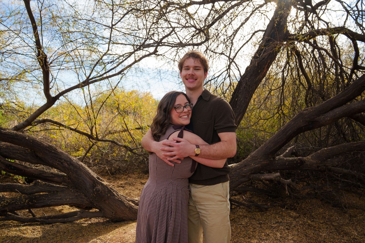 A smiling couple embraces outdoors among twisted desert trees with a bright sky backdrop.