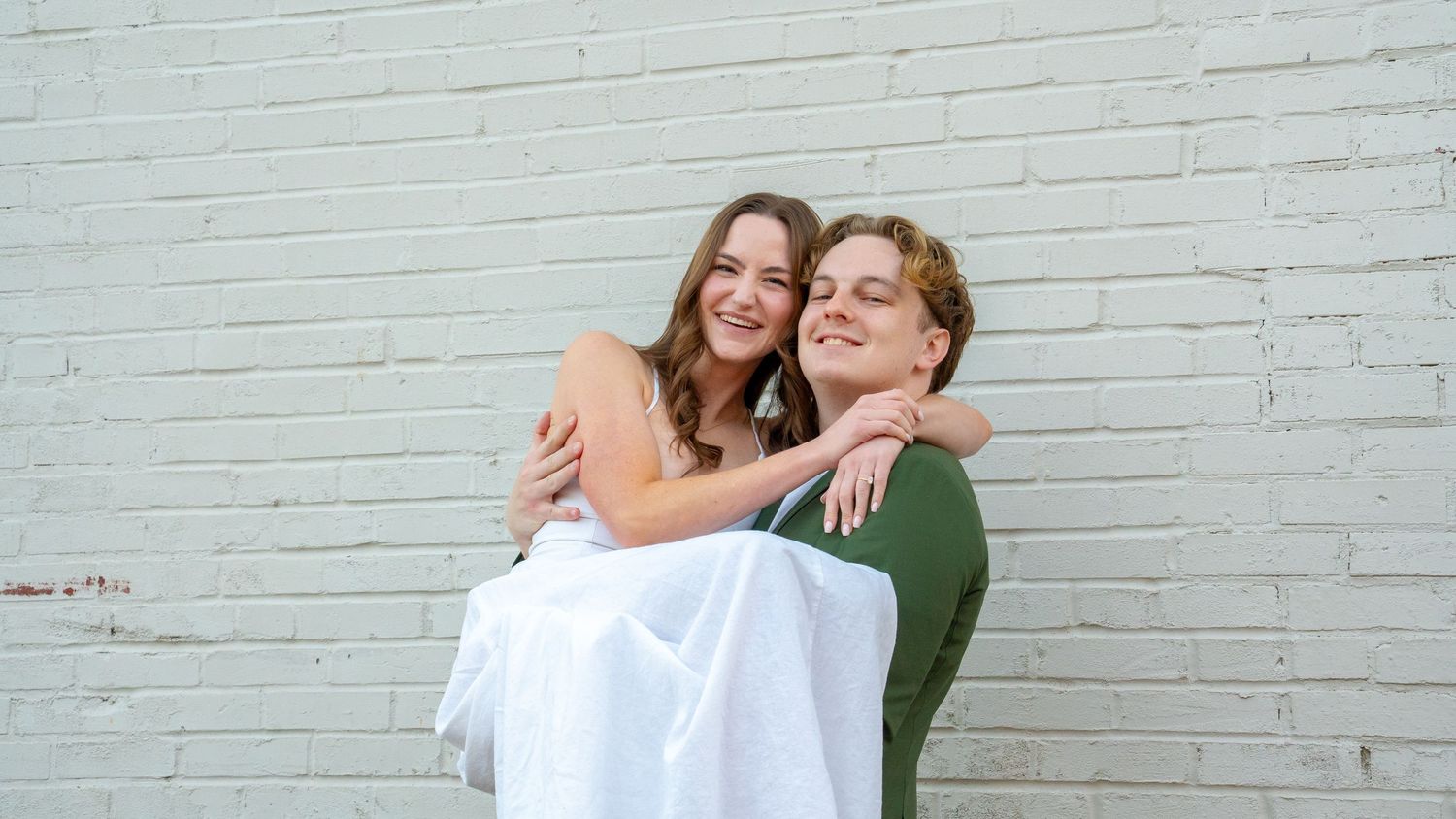 Couple in contrasting outfits perform playful dance moves against white brick background.