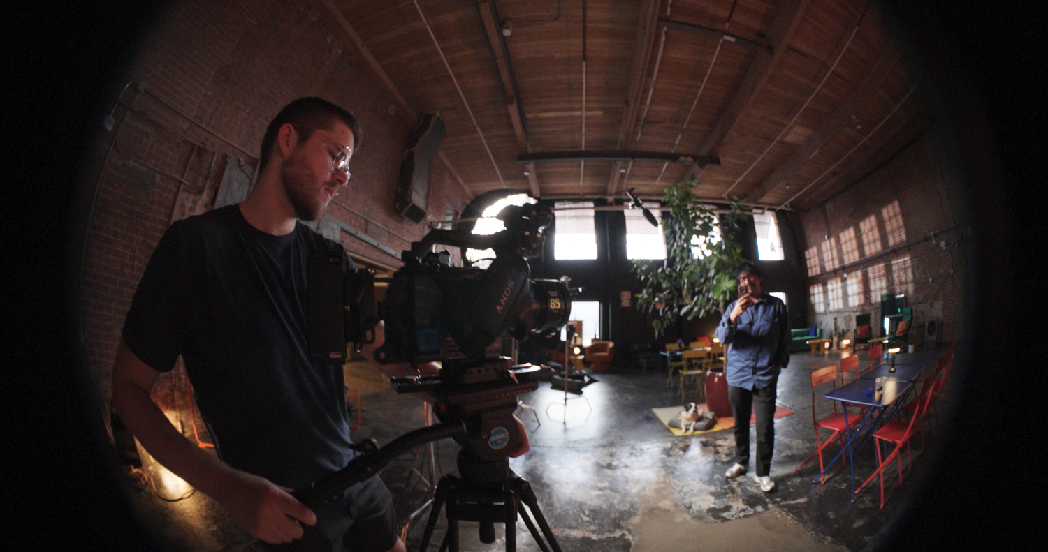 Fisheye lens view of a film crew in a brick studio, with a cameraman and subject during a video shoot.
