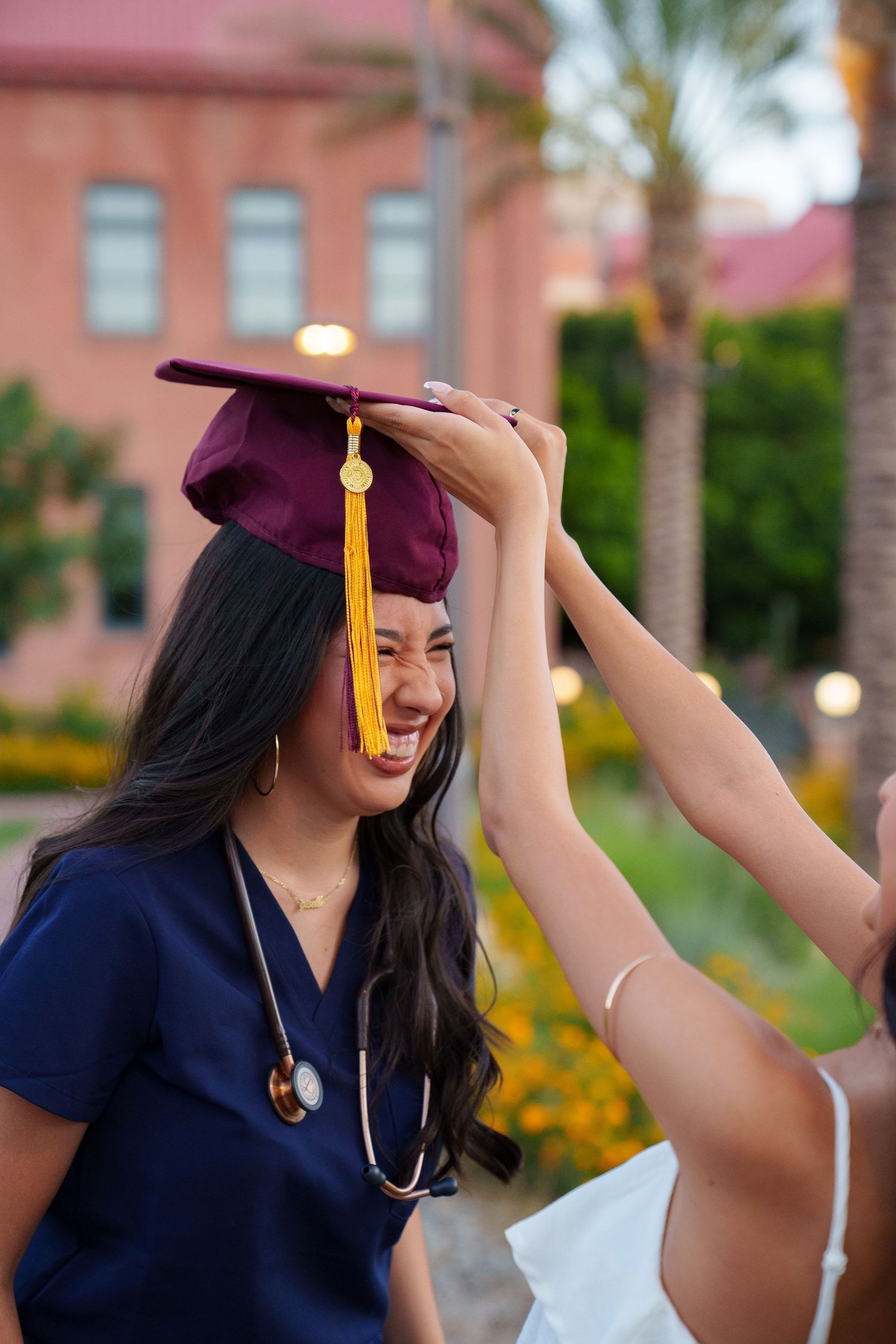 A joyful woman in nursing scrubs laughs as someone places a maroon graduation cap on her head outdoors.