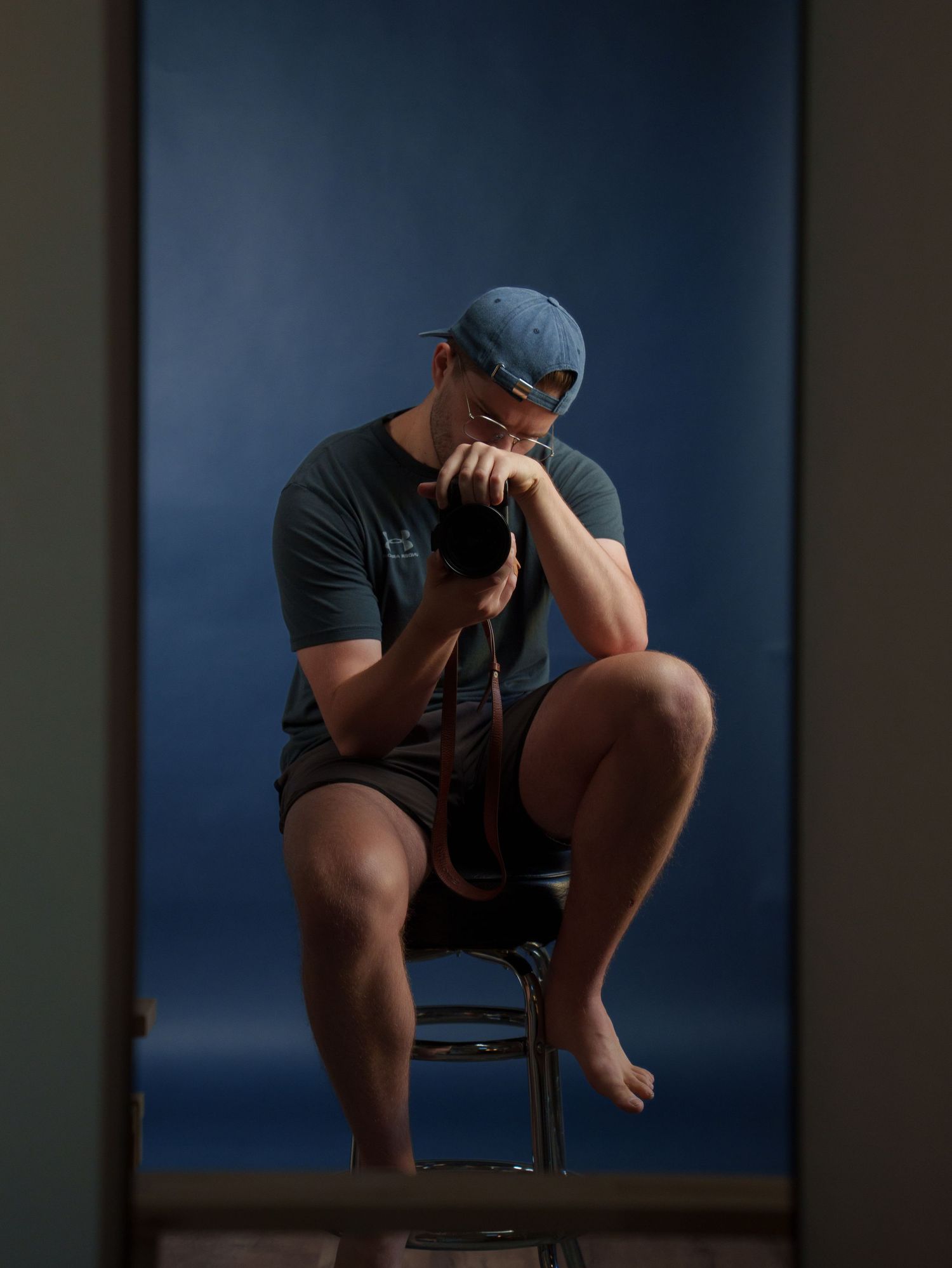 A man in a blue cap and shorts sits on a stool, holding a camera to his face against a blue backdrop.