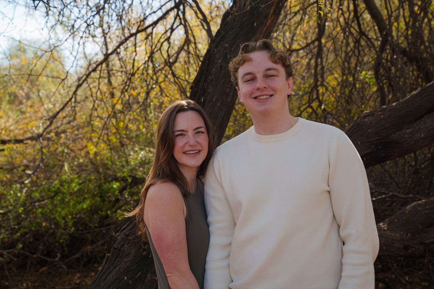 Two smiling young adults pose together outdoors among autumn trees with golden foliage in a natural setting.