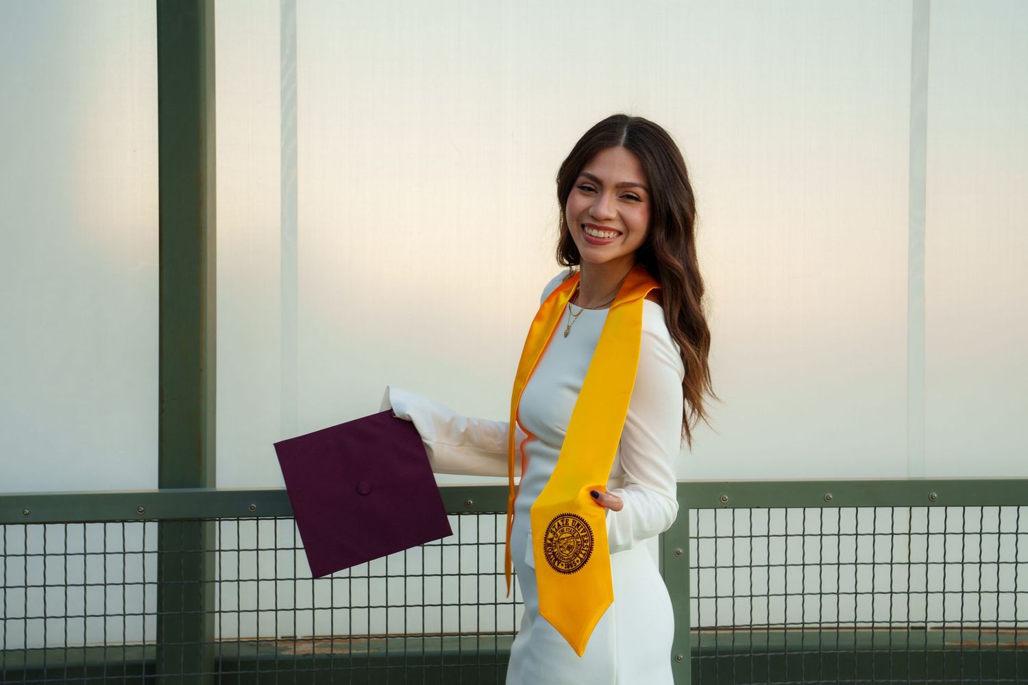 A graduate in a yellow and white outfit poses confidently against a bright outdoor background with railing.