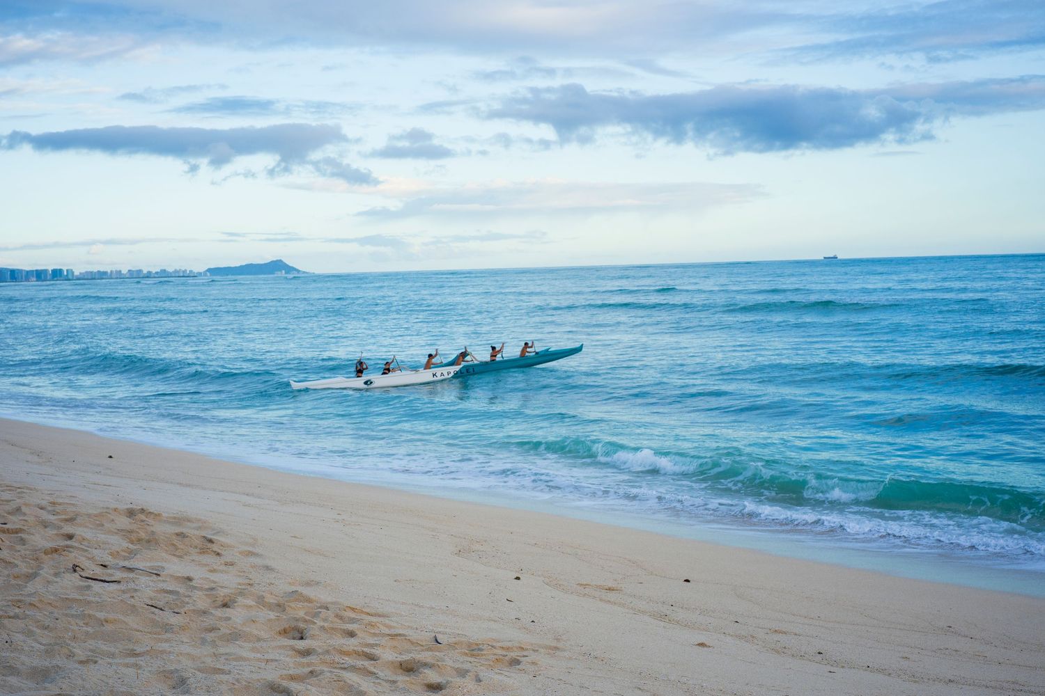 Panoramic view of a serene beach with blue ocean waves, cloudy sky, and a traditional Hawaiian rowing boat.