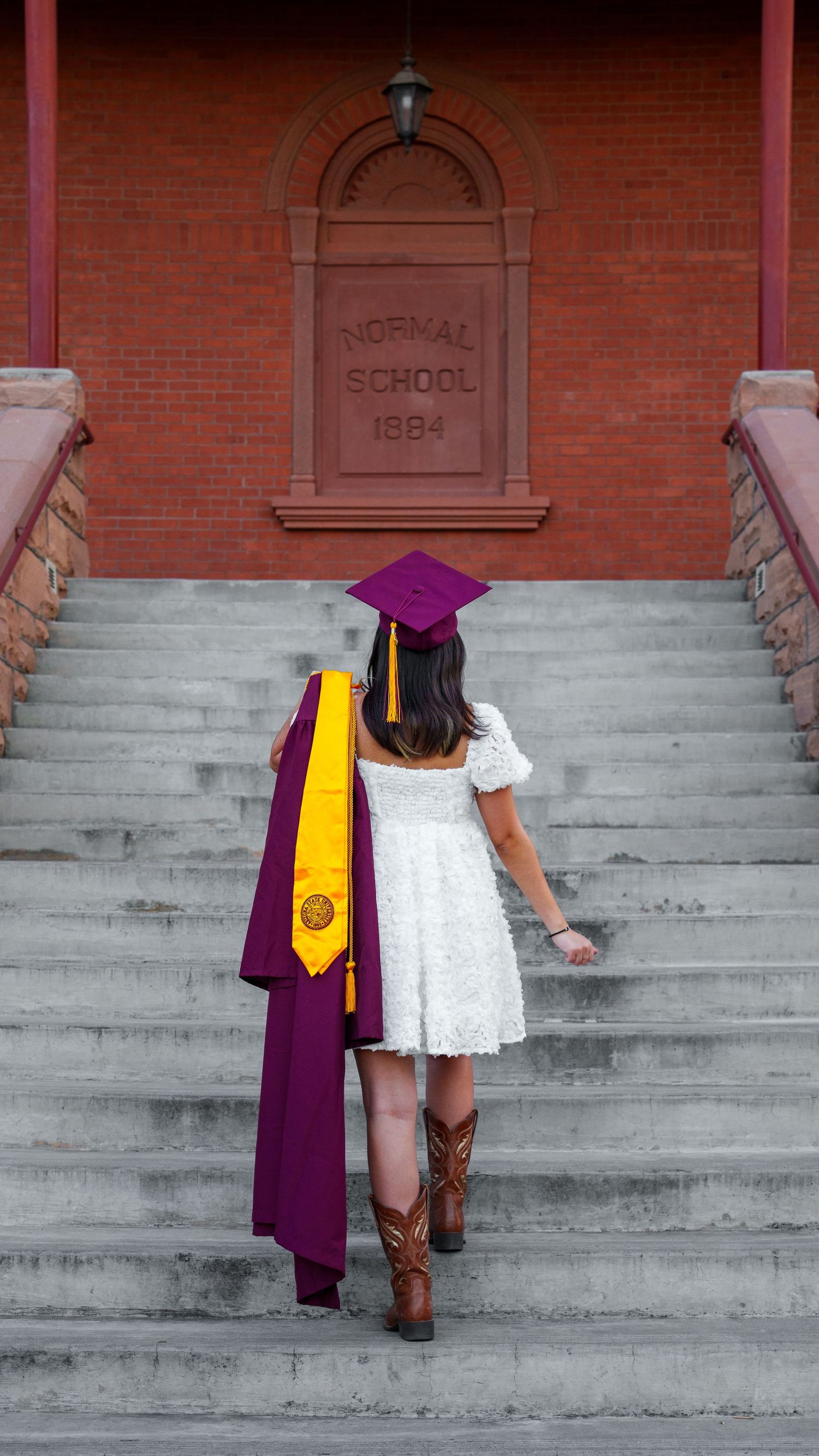 Graduate in white dress and purple graduation gown with cowboy boots stands on steps of historic Normal School building.