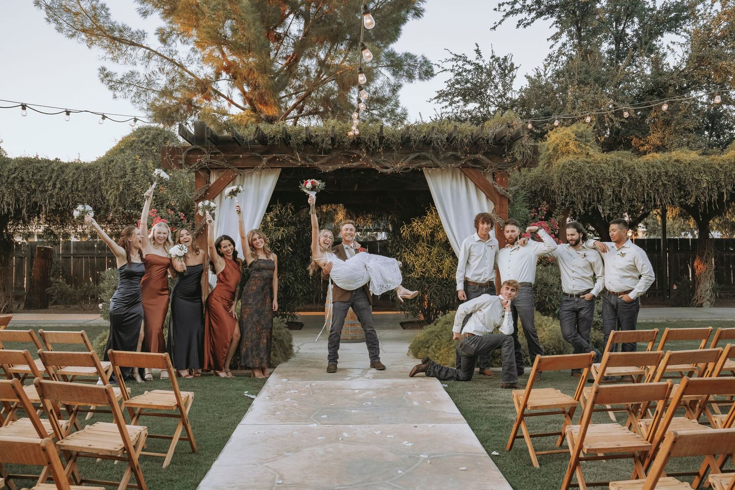 A wedding party with wooden folding chairs arranged outdoors for a rustic wedding ceremony with string lights overhead.