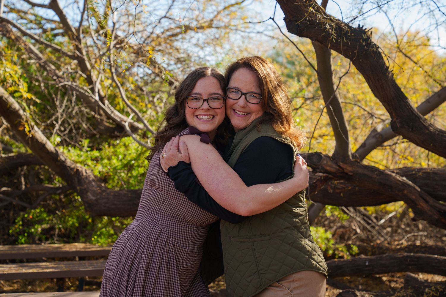 Two women with glasses hugging and smiling outdoors among autumn trees in a warm, joyful embrace.