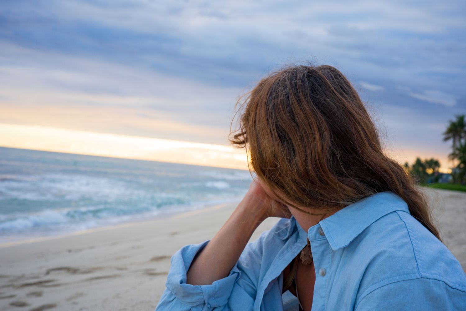 Person in blue shirt looking out at ocean waves during sunset.