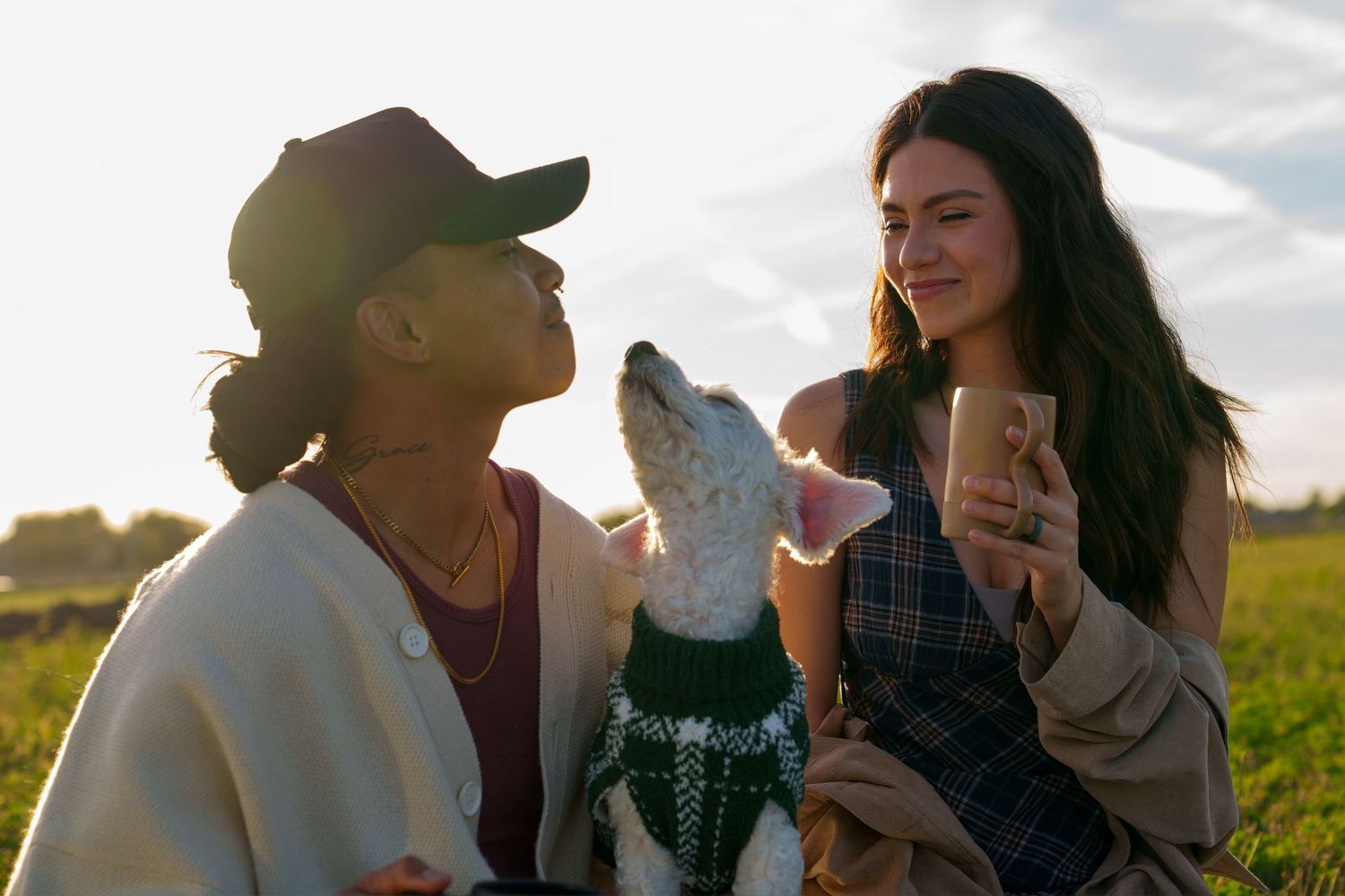Two women sitting outdoors with a dog in a sweater, sharing a warm moment in a sunlit green field.