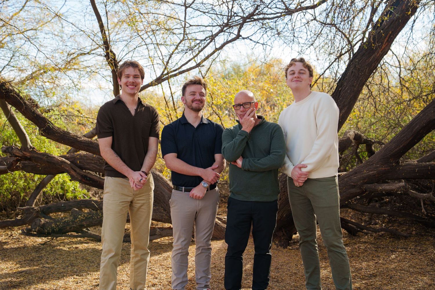 Four men smiling and posing together outdoors among desert trees on a sunny day.