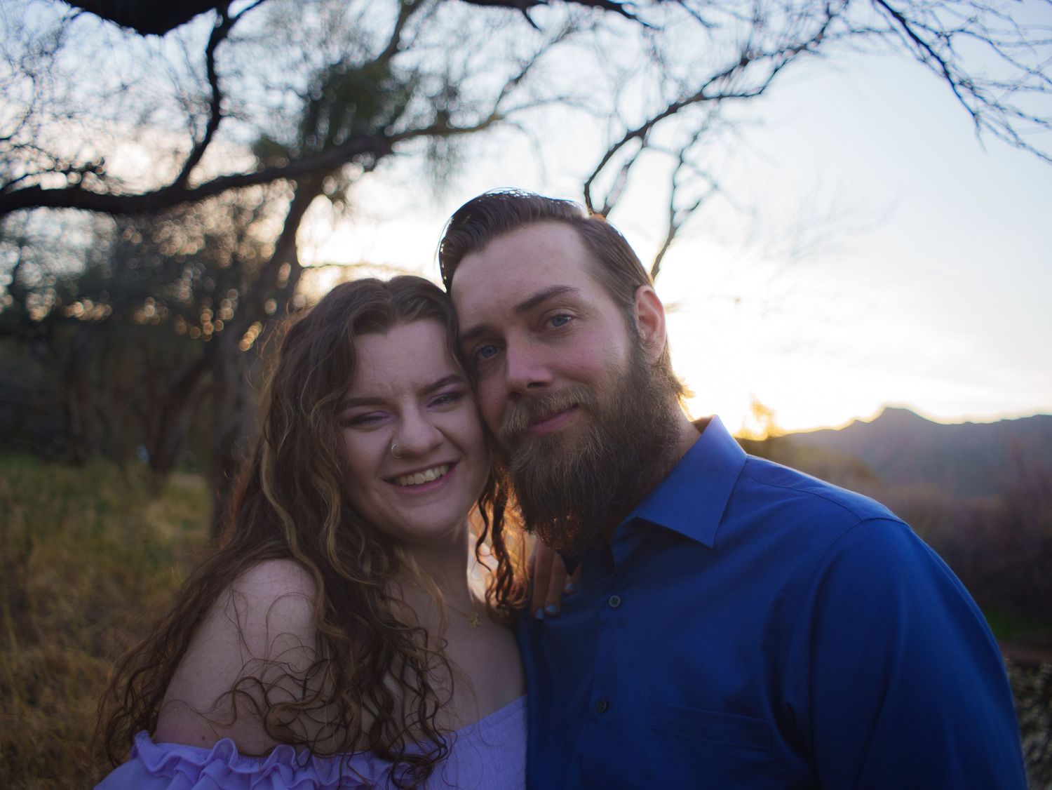 A couple poses together outdoors at sunset with trees and mountains visible in the background.