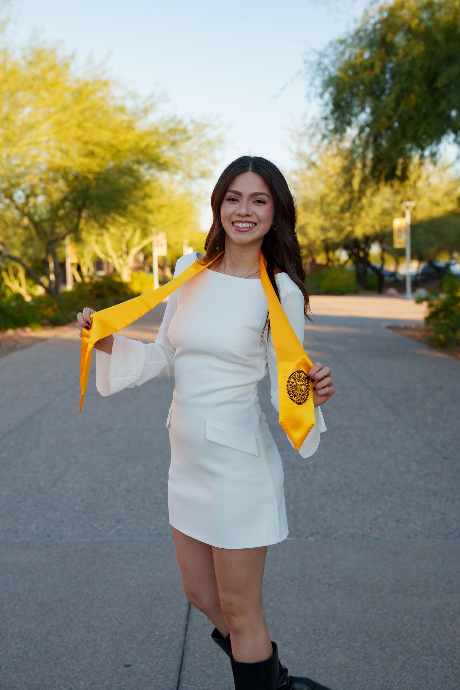 A person in a white dress holds up a yellow graduation stole while posing outdoors on a path with trees in the background.