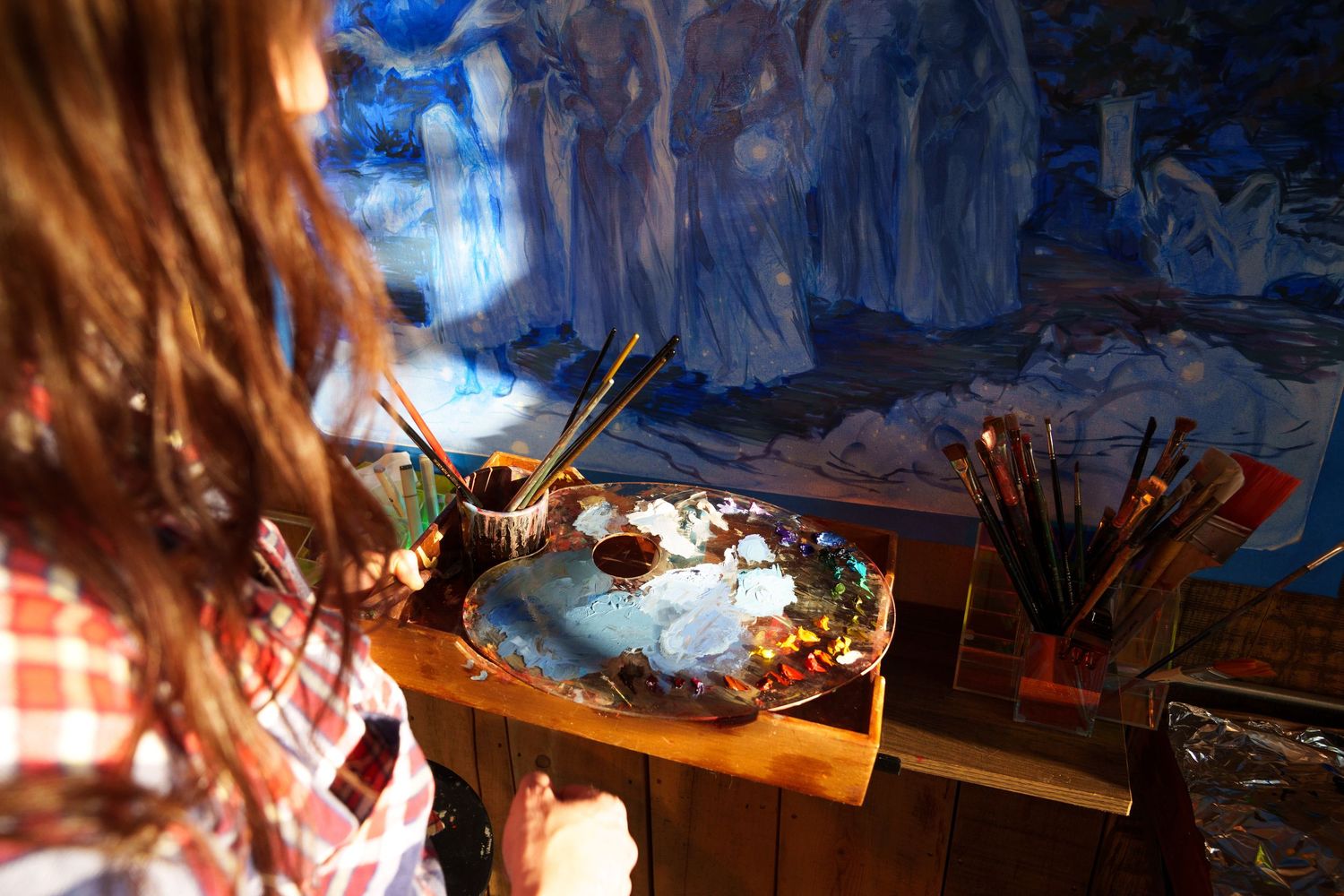 Artist painting a large blue fantasy mural with a wooden palette and brushes in a sunlit studio.