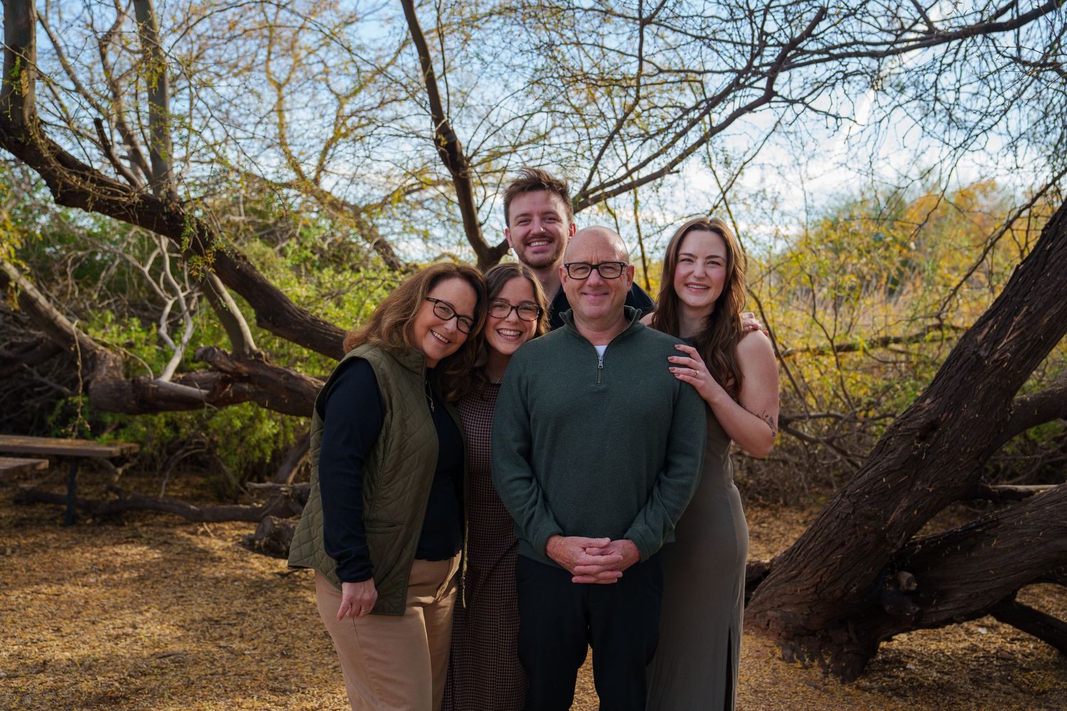 Family of five smiling together outdoors among trees in a natural setting during a fall family portrait session.