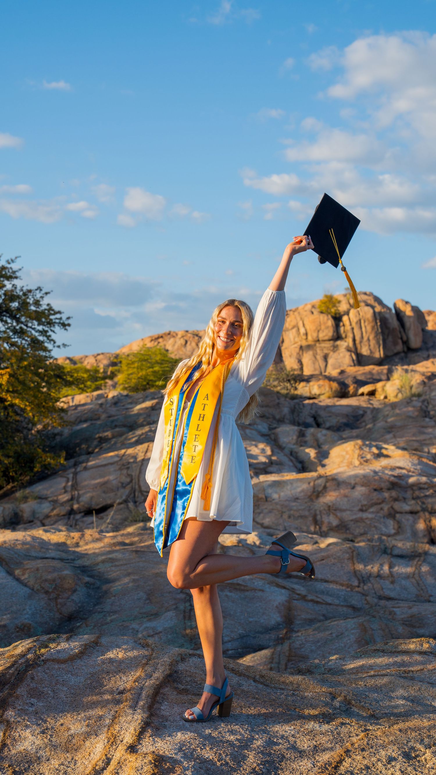 Graduate in cap and gown poses triumphantly on rocky terrain during golden hour sunset.