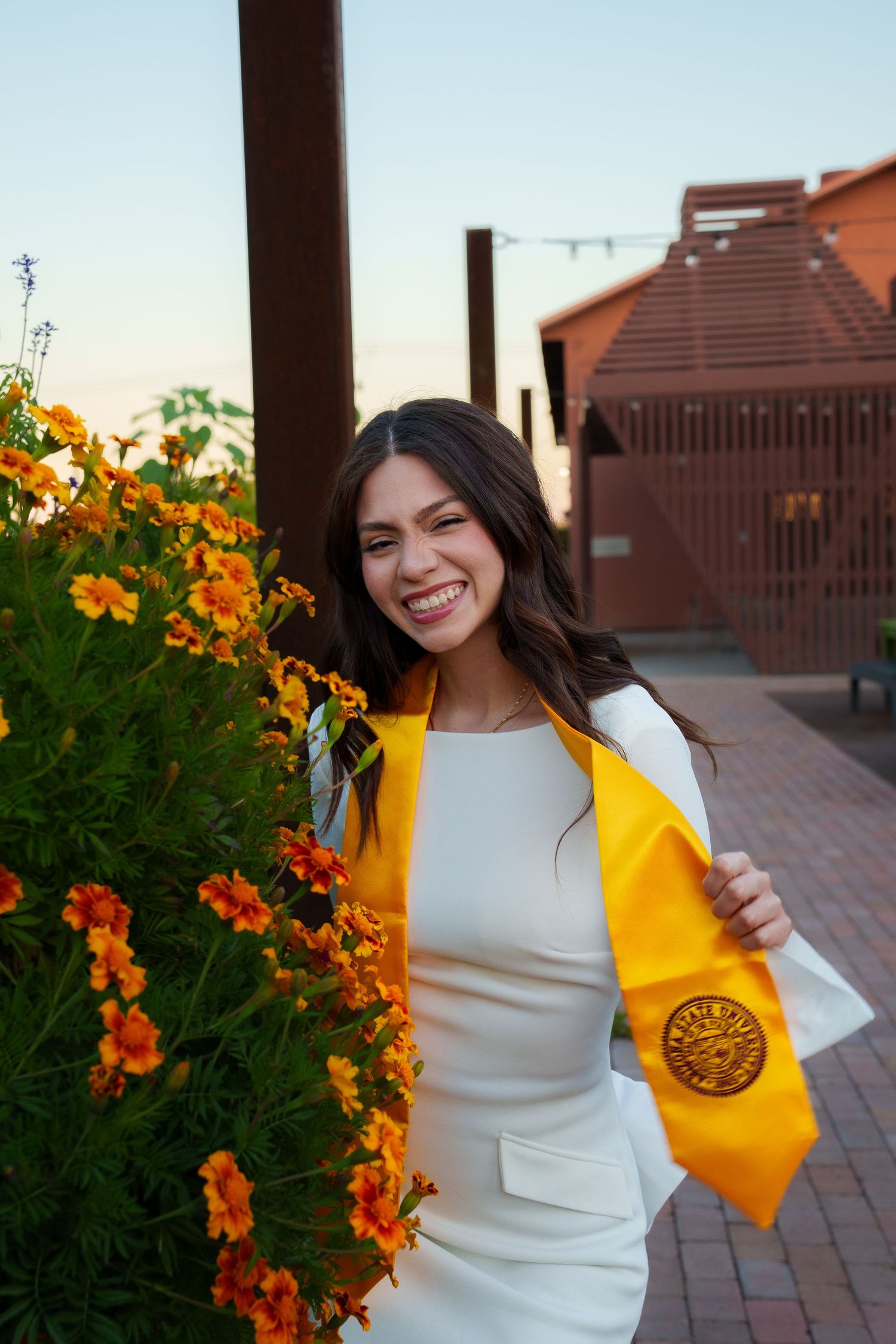 A graduate in a white dress and yellow graduation stole smiles joyfully next to vibrant orange flowers on a brick patio.