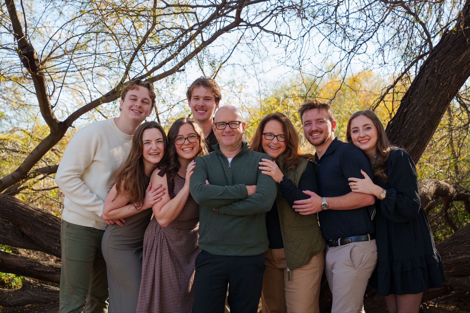 A large family of eight poses together outdoors under autumn trees, smiling warmly in coordinated earth-tone outfits.